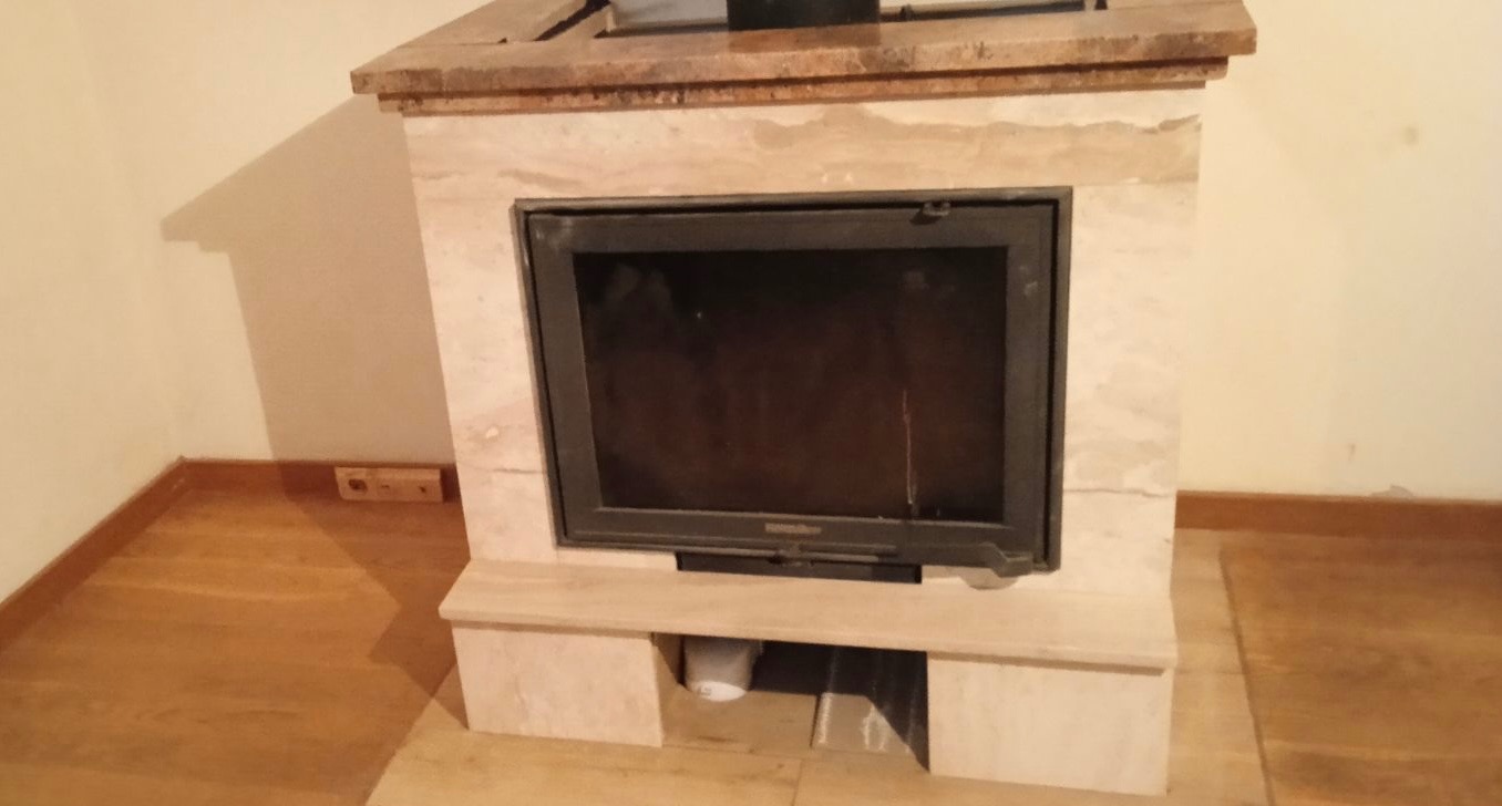 Obudowa kominka z jasnego kamienia z czarnym wkładem kominkowym, widoczna gładka powierzchnia kamienia i naturalne wzory, wnętrze kominka puste, na dole widoczne nieduże wgłębienie.