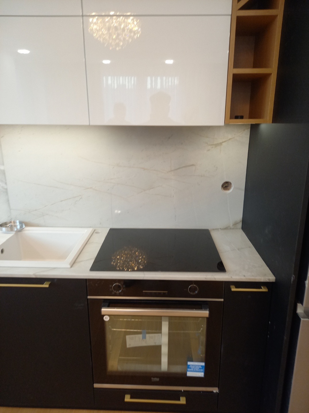 Nowoczesna kuchnia z czarnymi szafkami, złotymi uchwytami, białym blatem z marmurowym wzorem, płytą indukcyjną i piekarnikiem. Nad blatem białe szafki wiszące i marmurowa ściana. Widoczny fragment...