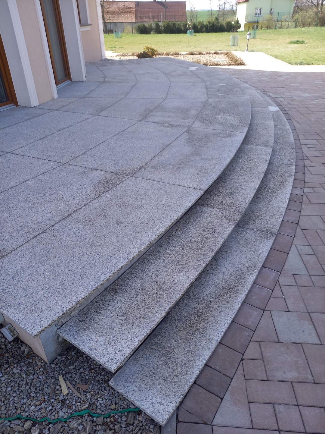 Trzy stopnie schodów zewnętrznych wyłożone szarą, ziarnistą płytą kamienną, prowadzące do tarasu z płyt tego samego materiału, obrzeże z kostki brukowej w odcieniach brązu, widok na dom i trawnik...