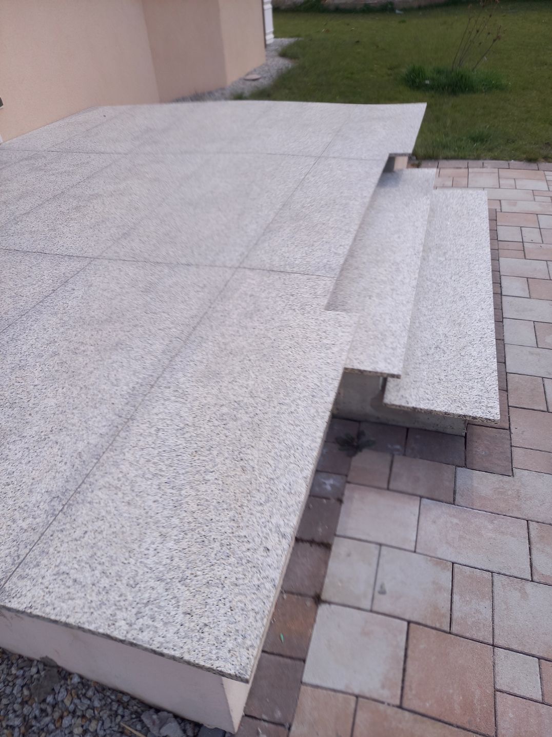 Granitowe schody wejściowe z tarasem, wyłożone jasnoszarą kostką, widok z boku na tle trawnika i elewacji budynku.