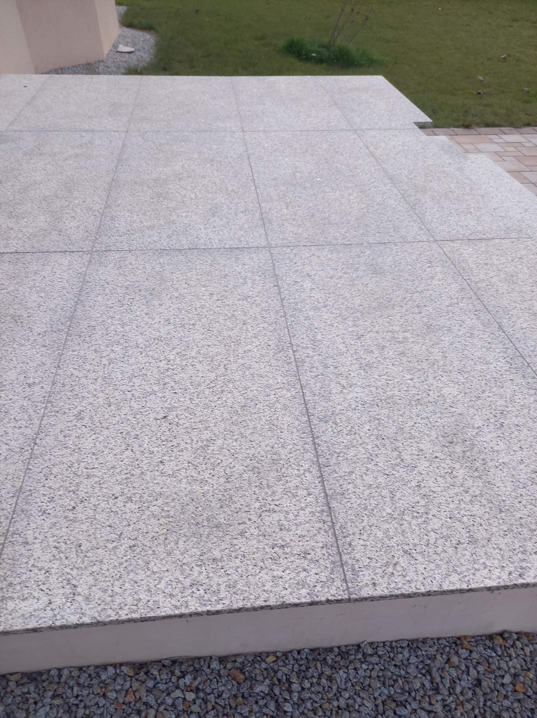 Jasnoszary taras wyłożony kwadratowymi płytami z drobnoziarnistego kamienia, widok z góry na fragment ogrodu z trawnikiem w tle.