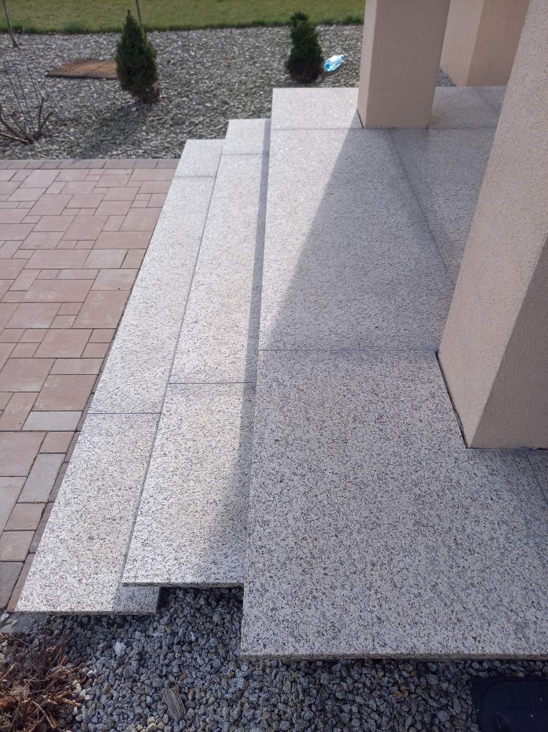 Zewnętrzne schody z jasnoszarego granitu prowadzące do wejścia, z widocznymi słupami podtrzymującymi zadaszenie, obok ścieżka z czerwonej kostki brukowej i ozdobny żwir.