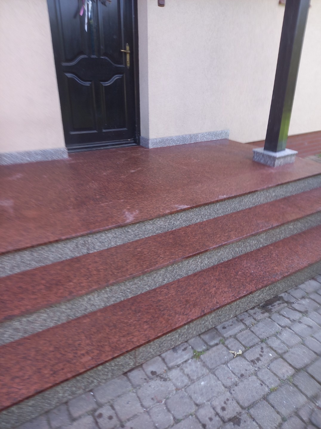 Granitowe schody zewnętrzne z czerwoną okładziną, prowadzące do ciemnych drzwi wejściowych. Wykończenie z szarego kamienia. Widok z dołu.