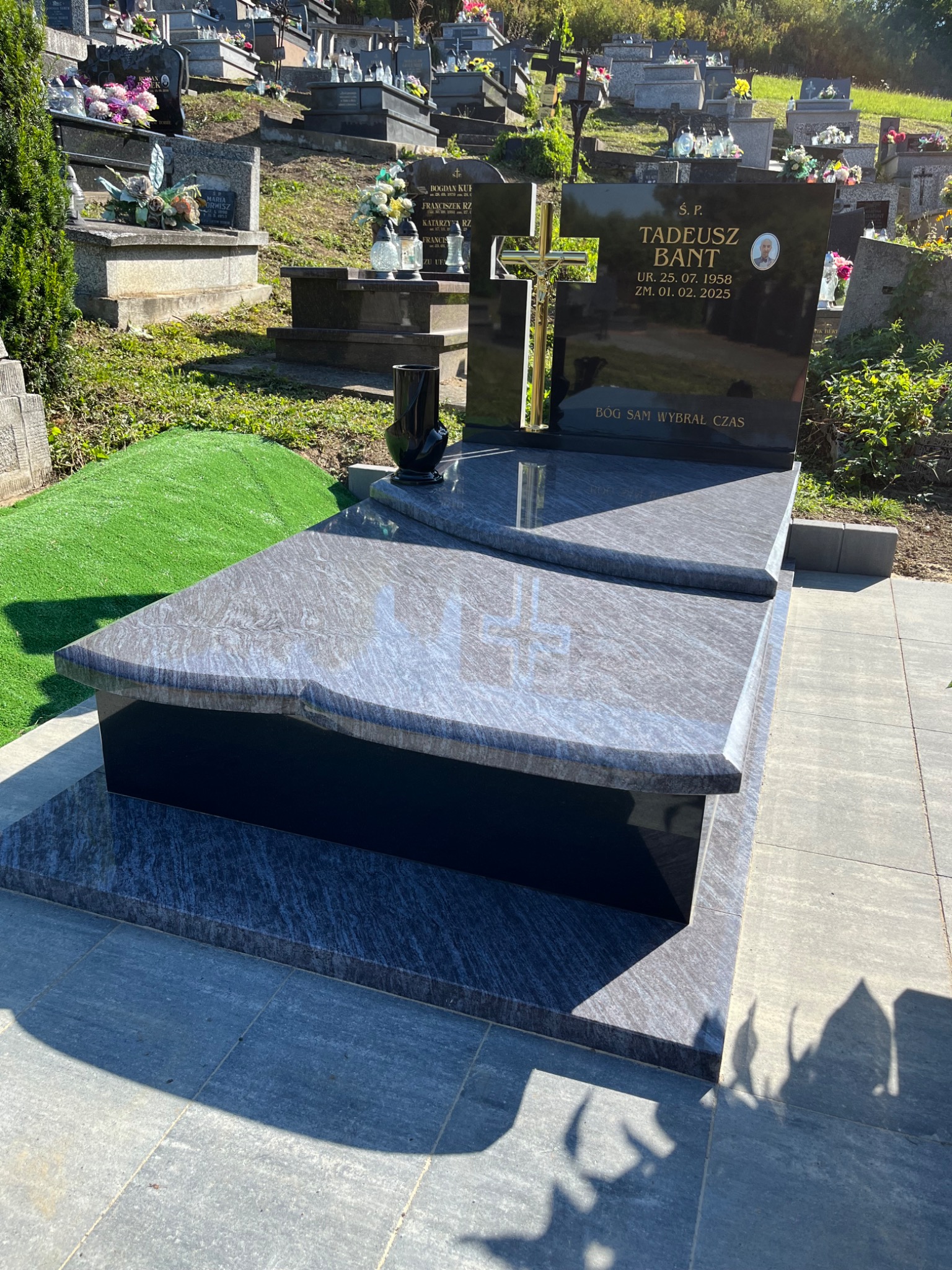 Granitowy nagrobek z czarną tablicą i złotym krzyżem na cmentarzu, w tle inne groby. Widoczna sztuczna trawa i kamienna podstawa nagrobka.