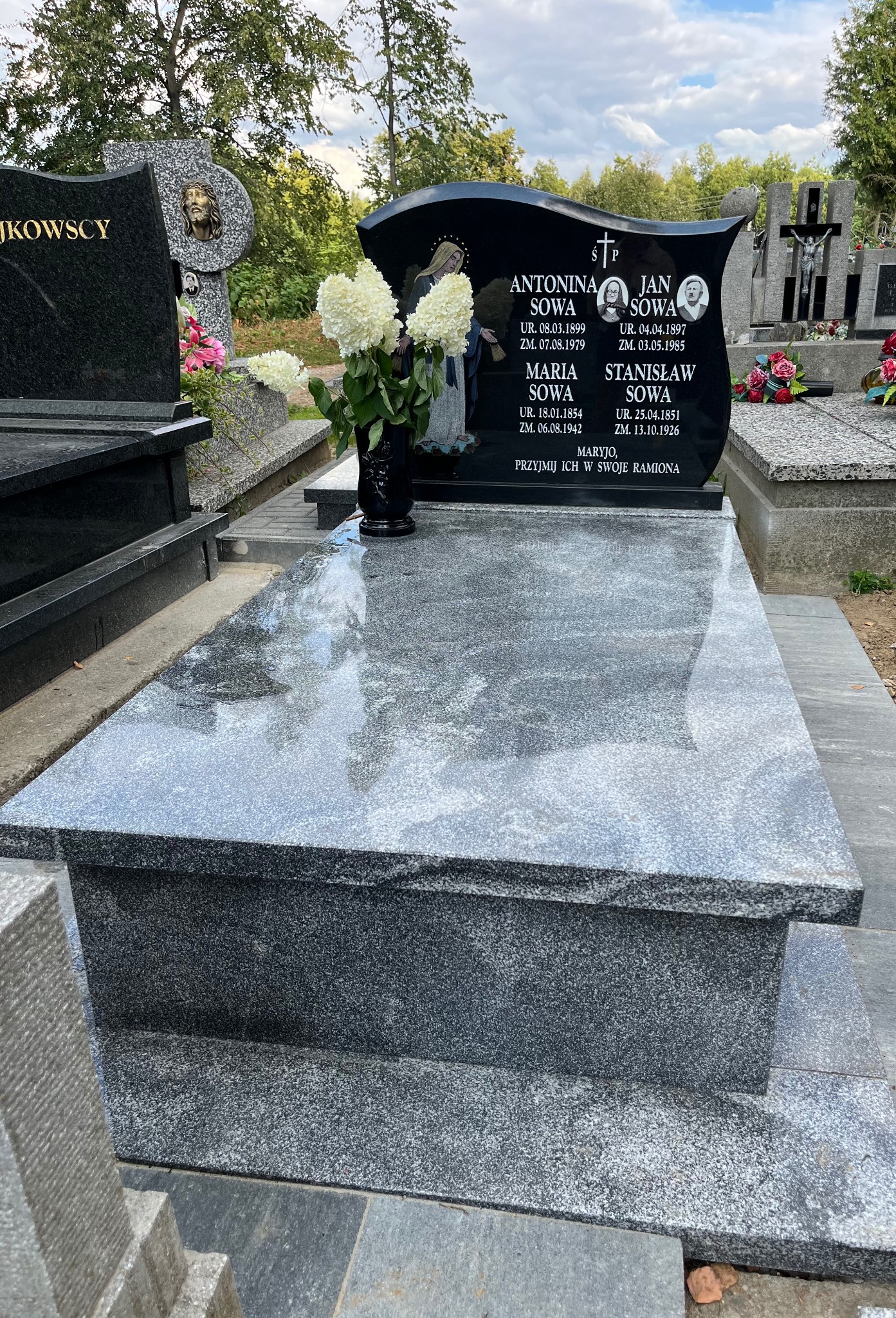 Granitowy nagrobek z czarną tablicą, na której widnieją imiona i nazwiska zmarłych, ozdobiony wazonem z białymi kwiatami. Widoczne inne nagrobki w tle.