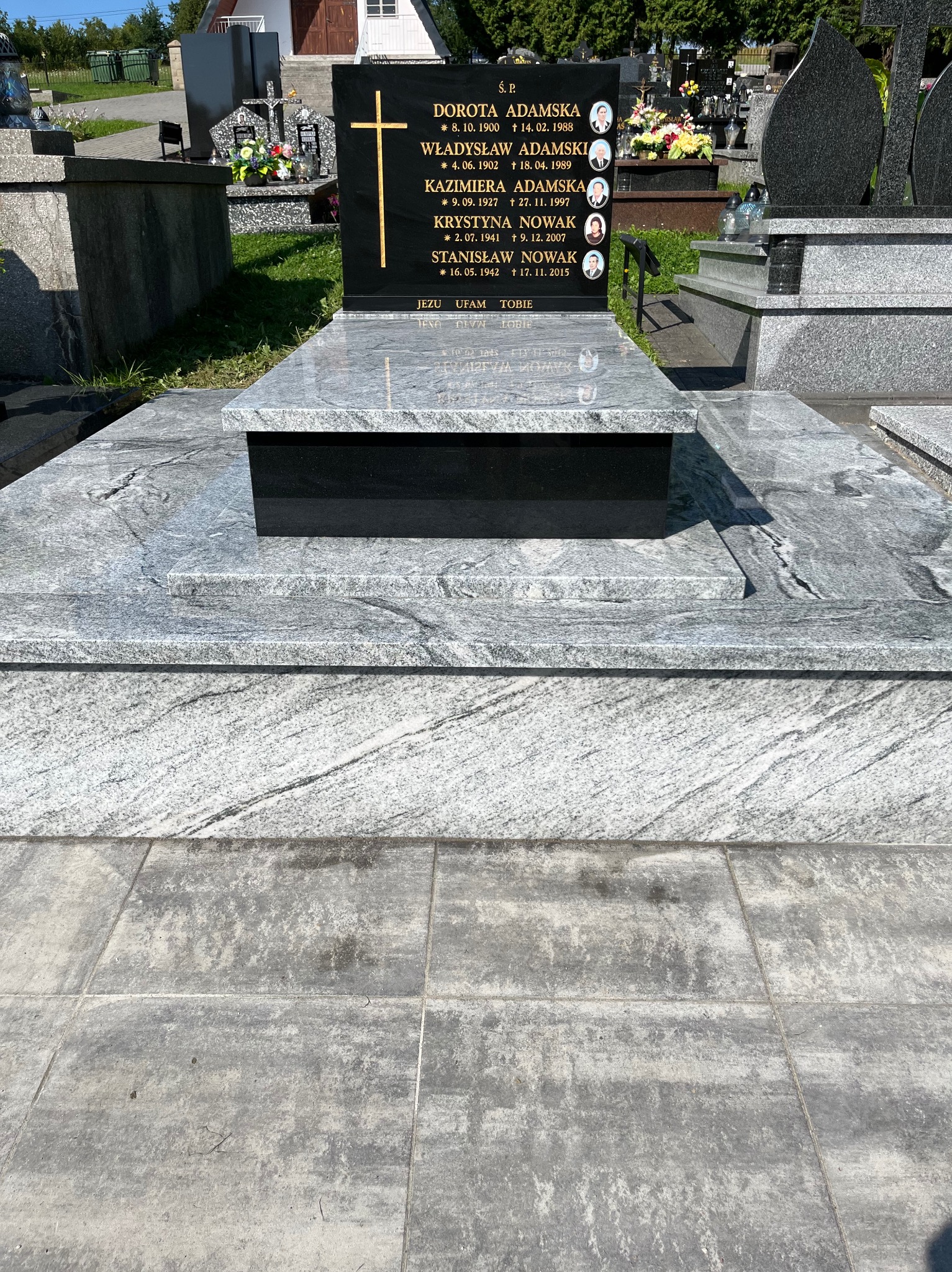 Szary nagrobek z czarną tablicą, złotym krzyżem i zdjęciami zmarłych na cmentarzu. Widoczne płyty nagrobne z szarego kamienia z wzorem.