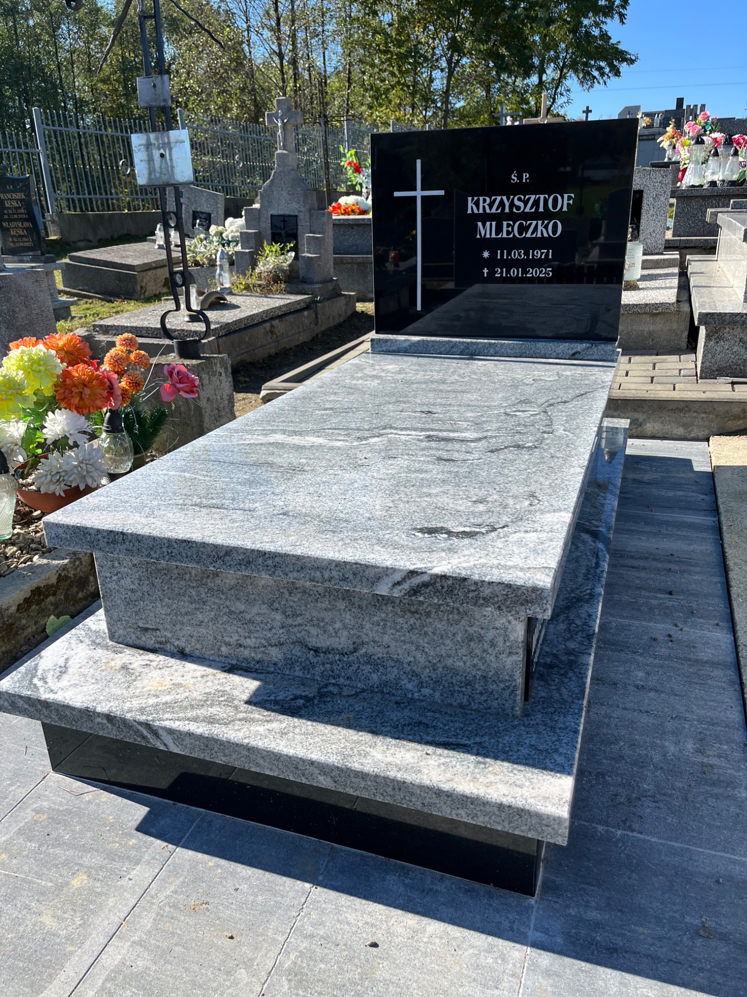 Granitowy nagrobek z czarną tablicą inskrypcyjną i kwiatami na cmentarzu w słoneczny dzień. Widoczne inne groby w tle.
