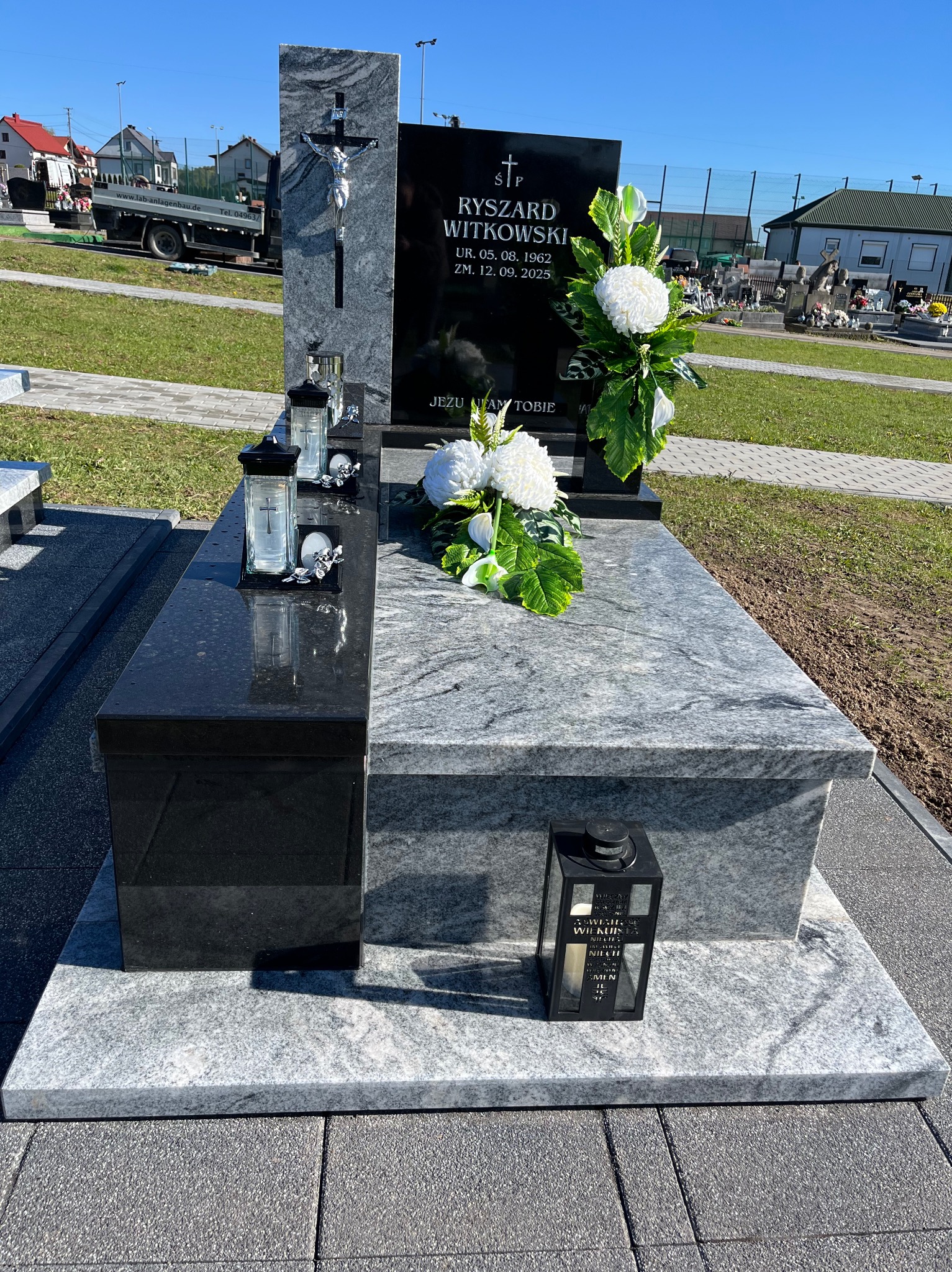 Granitowy nagrobek z czarną tablicą, białymi kwiatami i lampionami na cmentarzu. Widoczne nagrobki w tle, jasne oświetlenie słoneczne.