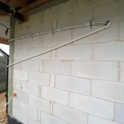 Biała ściana z betonu komórkowego z zamocowanymi rurami instalacyjnymi i przewodami elektrycznymi, widoczne elementy konstrukcji dachu.