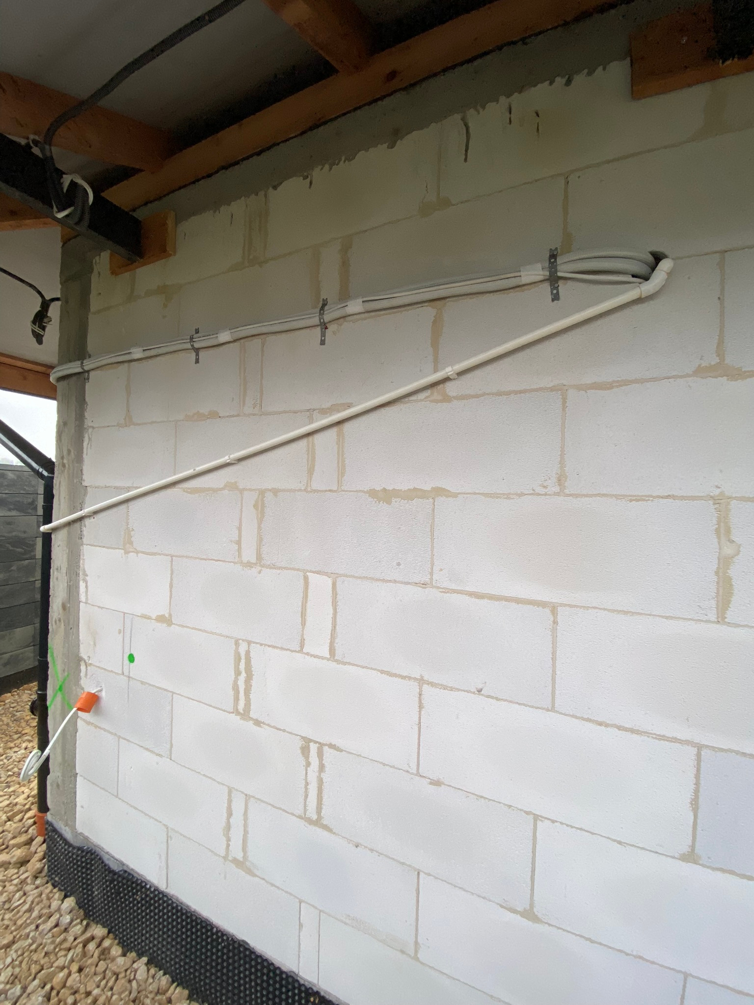 Biała ściana z betonu komórkowego z zamocowanymi rurami instalacyjnymi i przewodami elektrycznymi, widoczne elementy konstrukcji dachu.