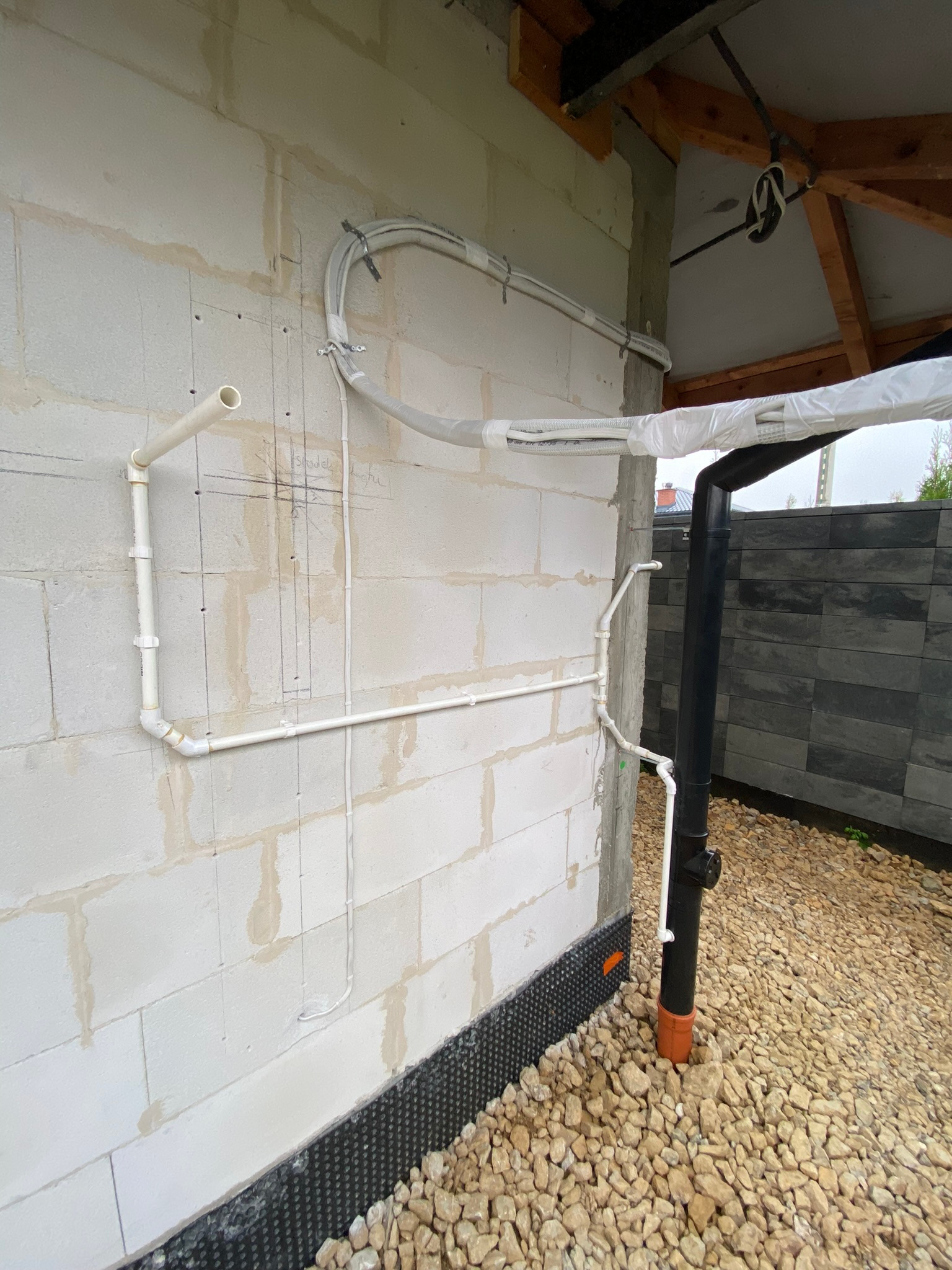 Biała ściana z bloczków z widocznymi instalacjami: rurami PCV, przewodami w białej osłonie i czarną rurą spustową. Widoczne są także zaznaczenia ołówkiem na ścianie i kamienny podkład.