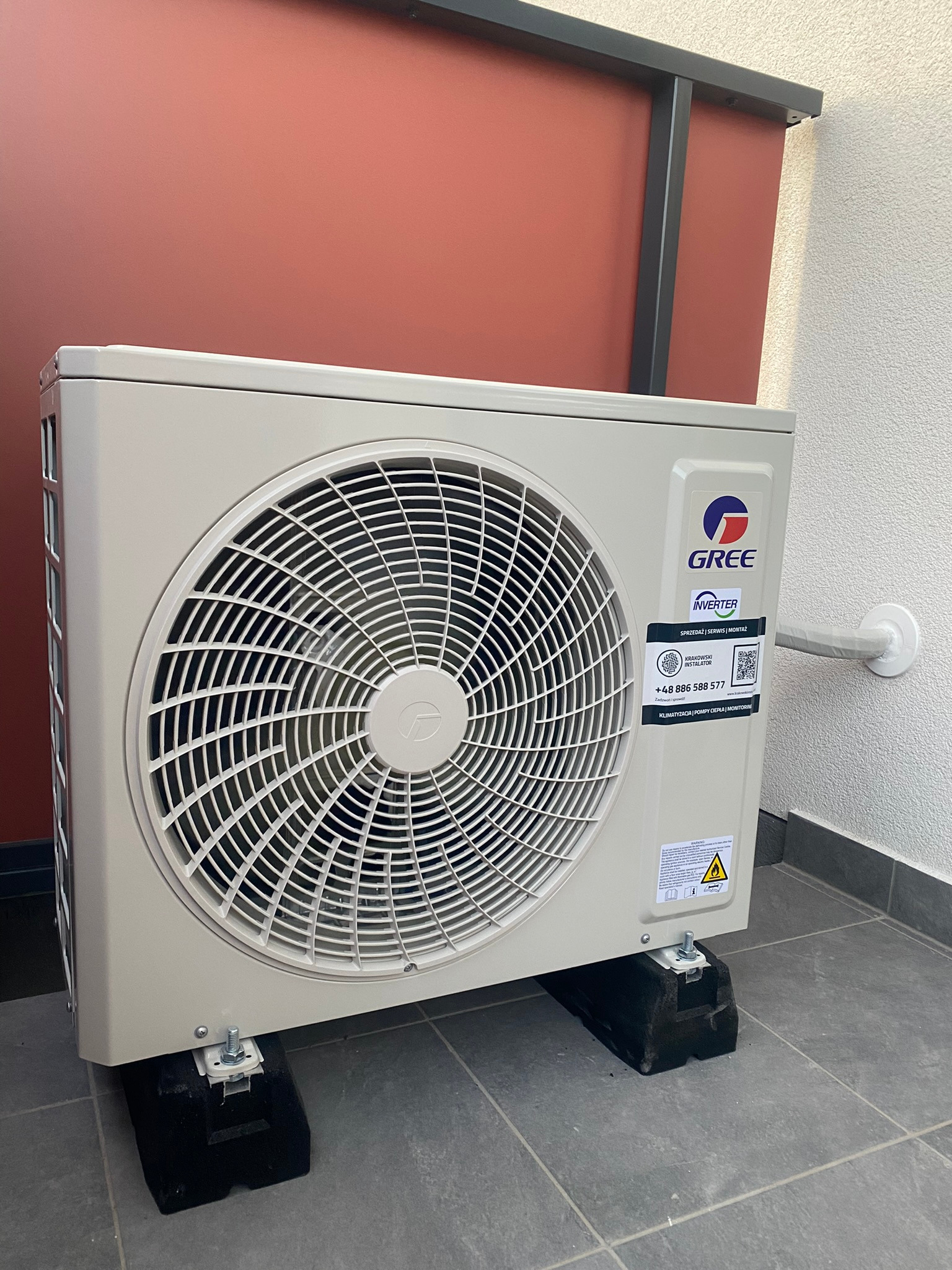 Zewnętrzna jednostka klimatyzacji marki Gree Inverter zamontowana na czarnych podkładkach antywibracyjnych, przy ścianie pomalowanej na terakotowy kolor, z widocznym przewodem instalacyjnym.