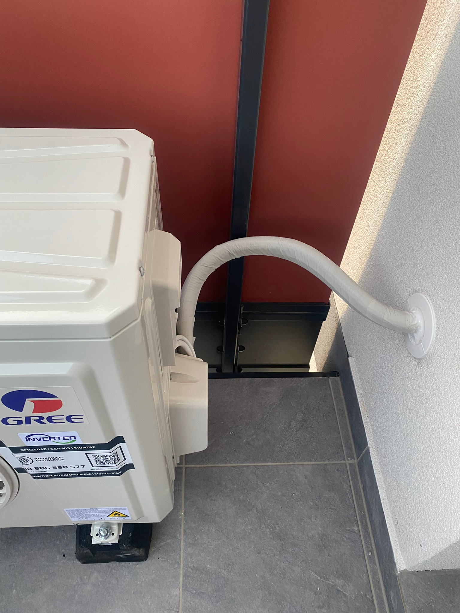 Zewnętrzna jednostka klimatyzacji marki Gree zamontowana przy ścianie budynku, z widocznym przewodem odprowadzającym zabezpieczonym białą izolacją.