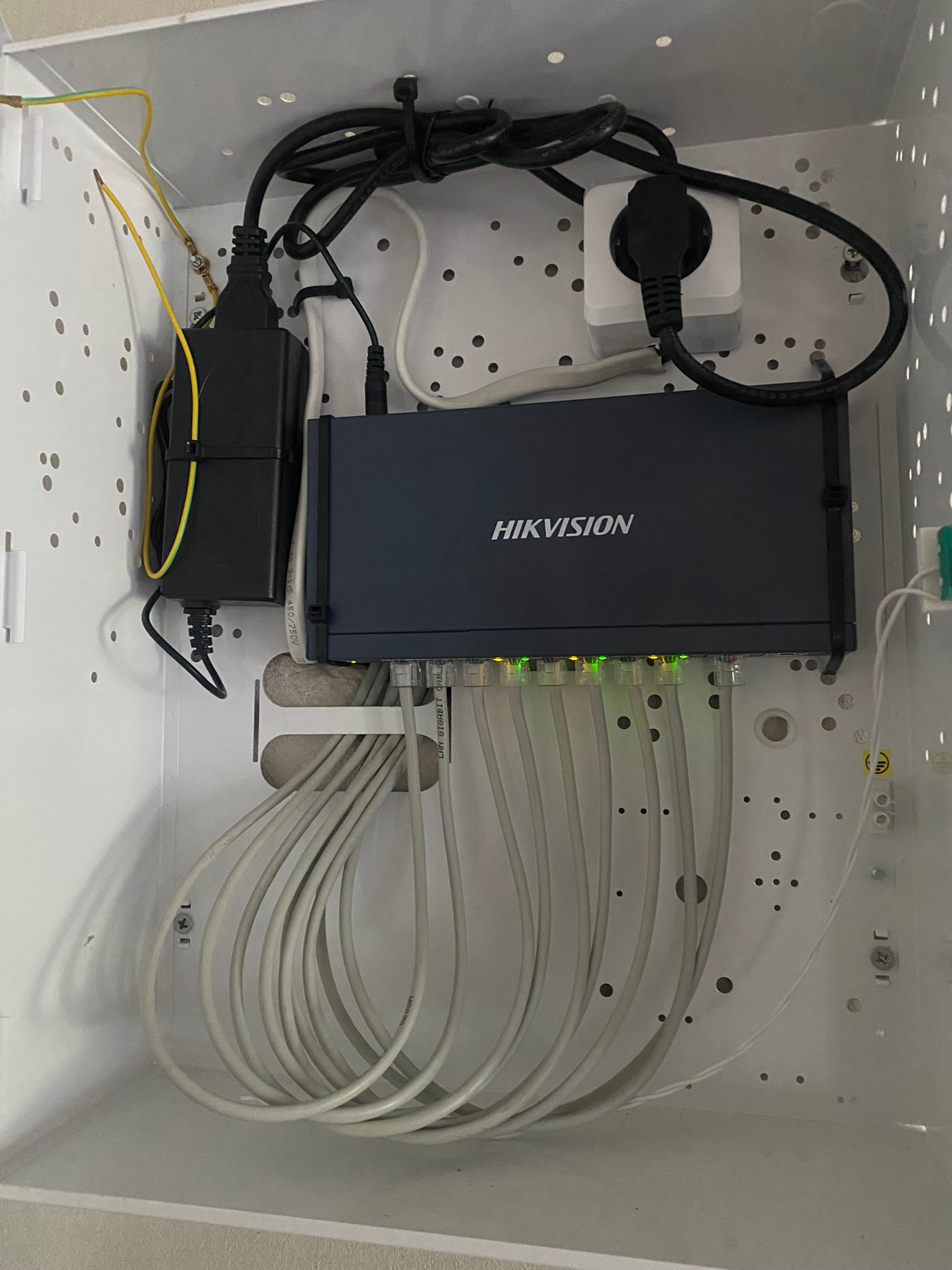 Zamknięta szafka z zamontowanym switchem Hikvision, podłączonymi kablami Ethernet i zasilaczem. Widoczne przewody zasilające i uziemiające. Kabel Ethernet zabezpieczony opaską zaciskową.