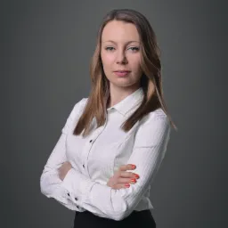 Portret kobiety z założonymi rękami, ubrana w białą koszulę z delikatnym wzorem na tle szarego tła.