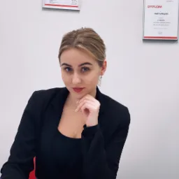 Kobieta w czarnej marynarce siedzi przy biurku, pisząc w otwartym notatniku; na ścianie wiszą dwa dyplomy, jeden z nich to dyplom III miejsca w konkursie sprzedaży APE/APE WN Agencji w 2019 roku.