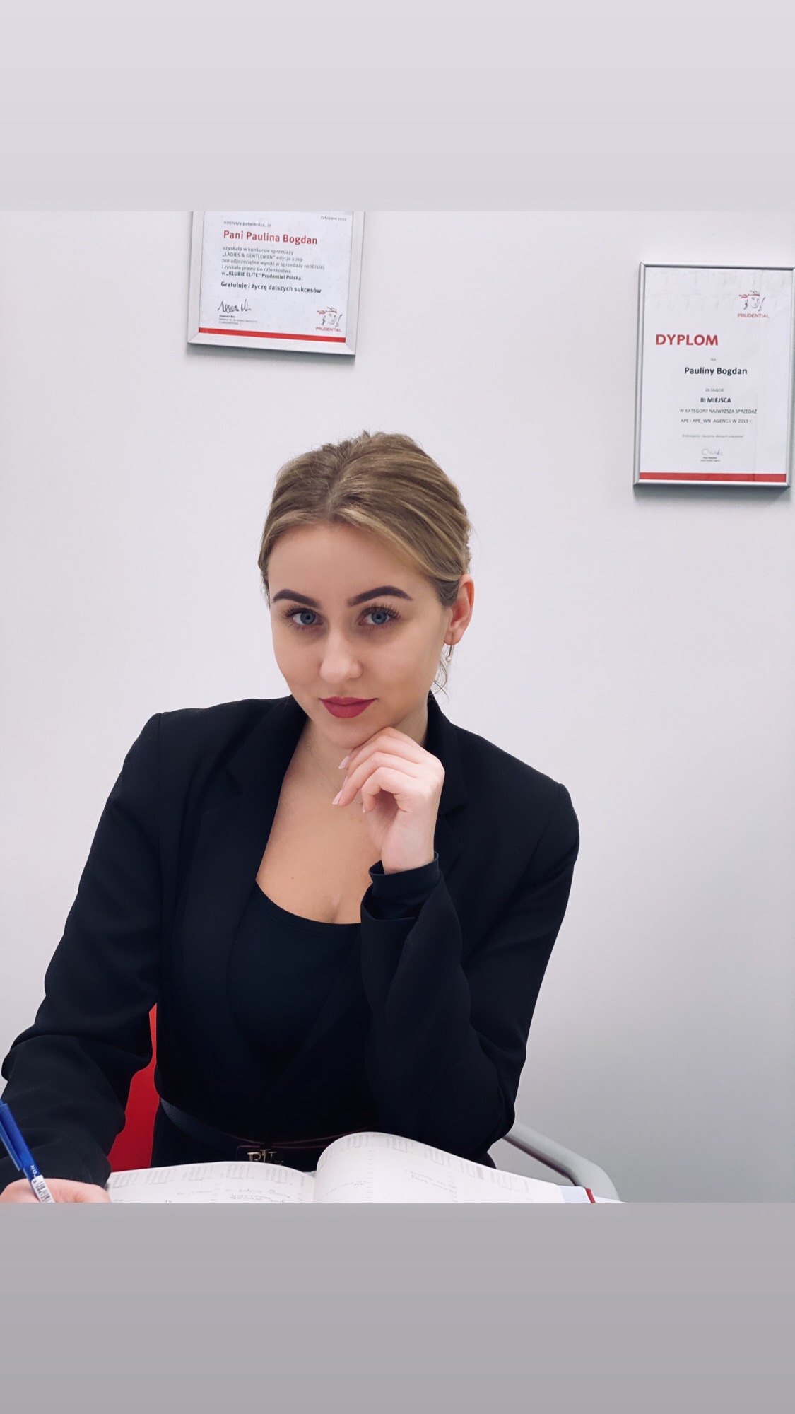 Kobieta w czarnej marynarce siedzi przy biurku, pisząc w otwartym notatniku; na ścianie wiszą dwa dyplomy, jeden z nich to dyplom III miejsca w konkursie sprzedaży APE/APE WN Agencji w 2019 roku.