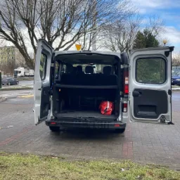 Srebrny van z otwartymi tylnymi drzwiami, widoczny pusty bagażnik z czerwoną torbą i trójkątem ostrzegawczym, zaparkowany na chodniku w miejskim otoczeniu.