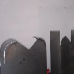 Dwa elementy metalowe, prawdopodobnie formy lub narzędzia, stojące przed białą ścianą. Jeden element ma zaokrąglony kształt z ostrym wycięciem, a drugi ma prostokątny kształt z wycięciem...