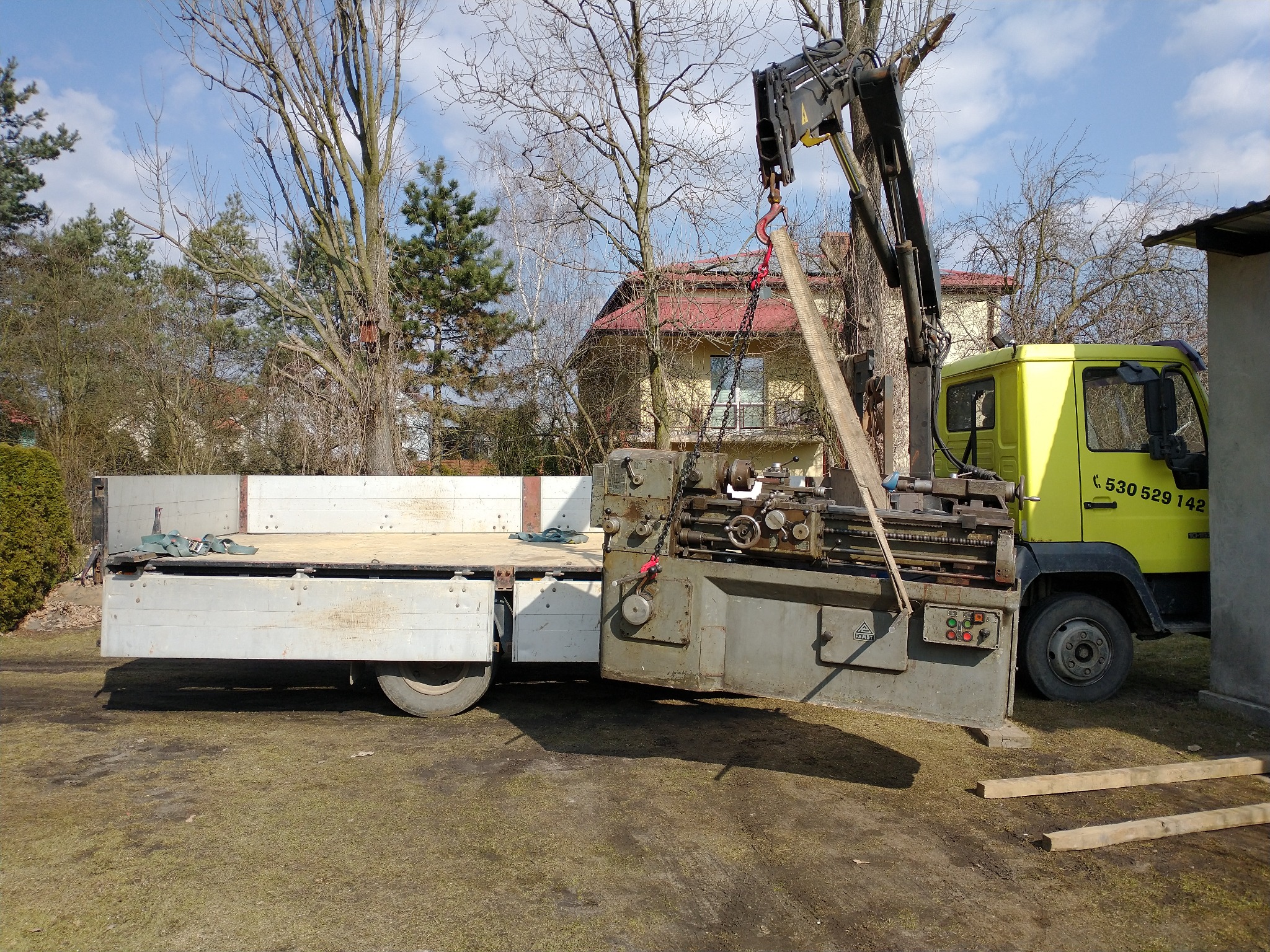 Żółty samochód ciężarowy z dźwigiem załadowuje tokarkę na platformę; widoczne drzewa i budynek w tle.