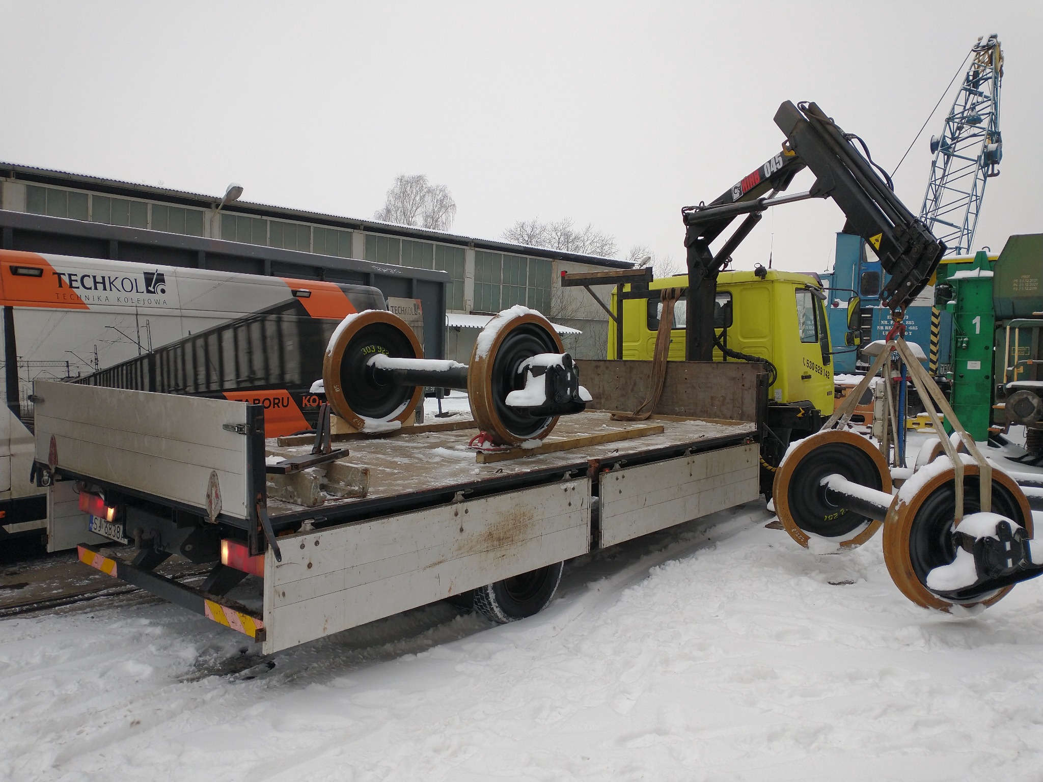 Załadunek zestawów kołowych do wagonów kolejowych na platformę ciężarówki z dźwigiem w zimowej scenerii.