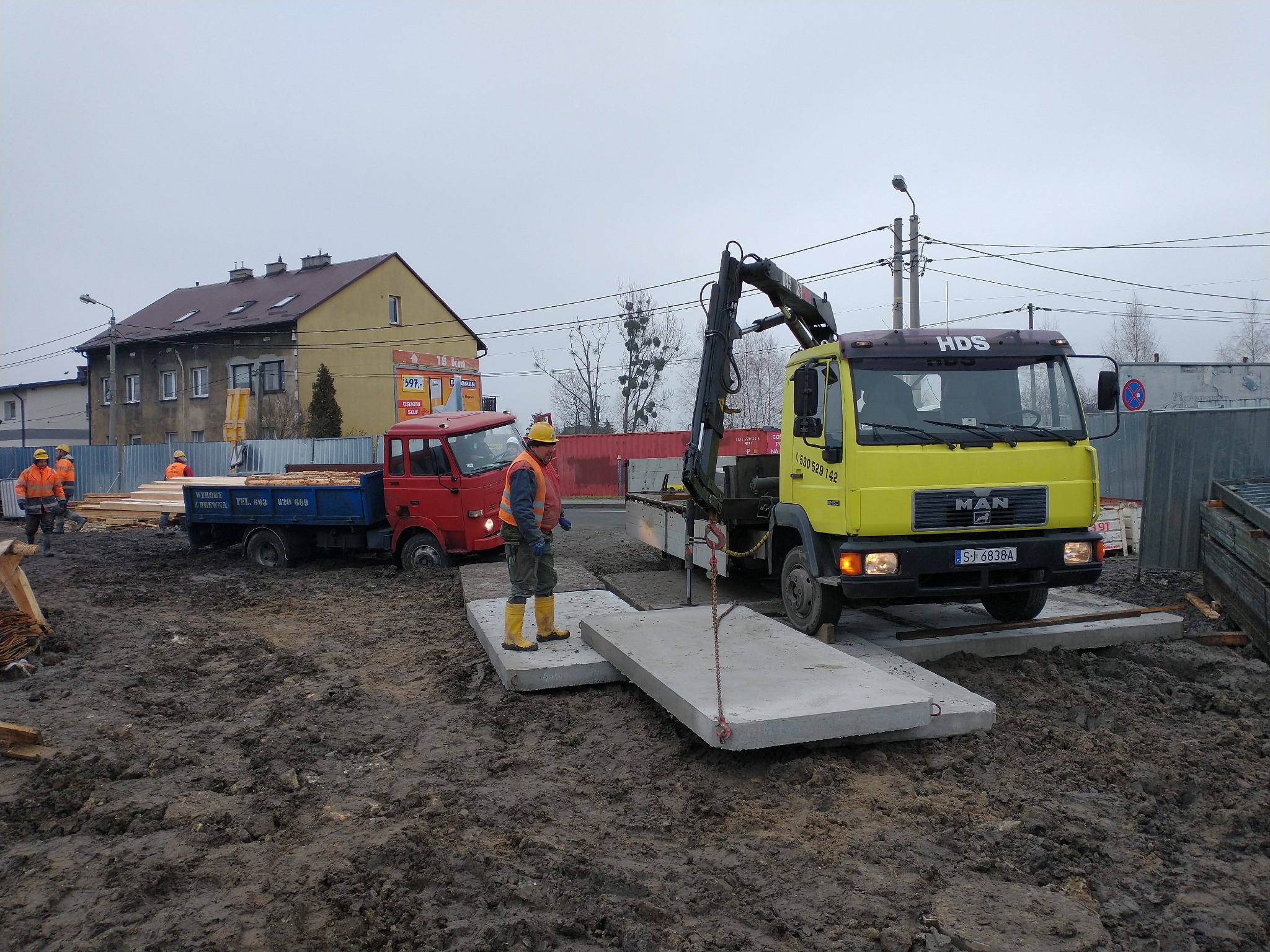 Żółty samochód ciężarowy MAN HDS z dźwigiem, podnosi betonową płytę na placu budowy, pracownik w żółtych butach nadzoruje proces, w tle czerwona ciężarówka z drewnem.