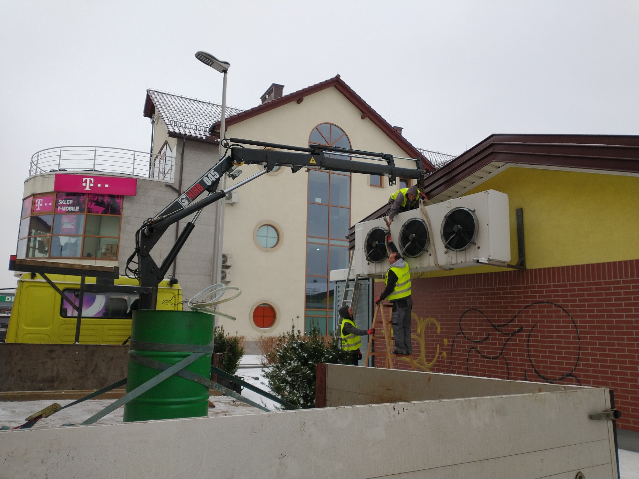 Montaż klimatyzacji przemysłowej na dachu budynku przez ekipę w kamizelkach odblaskowych z użyciem dźwigu zamontowanego na żółtej ciężarówce, widoczna zielona beczka w skrzyni ładunkowej.