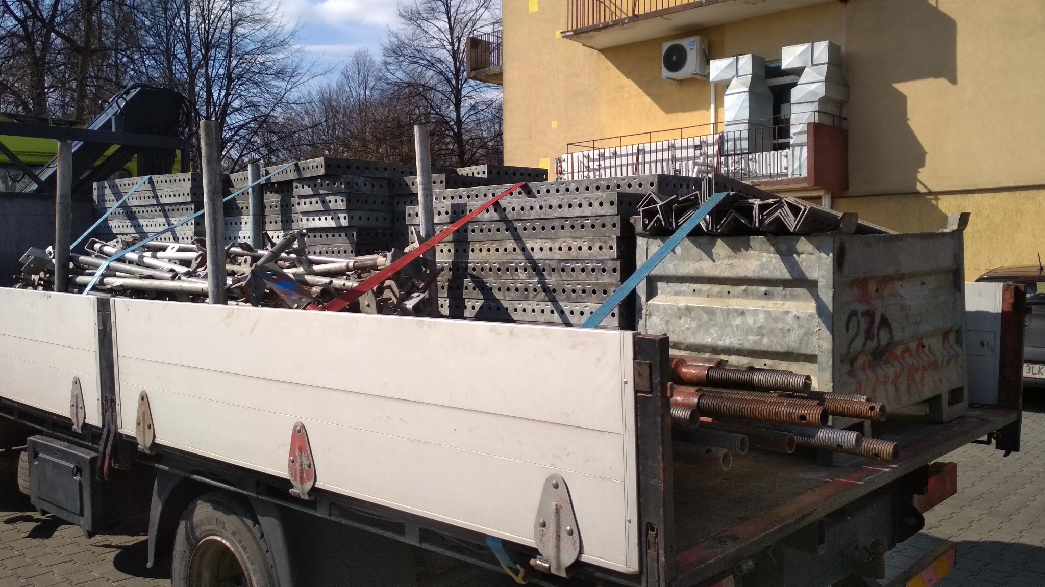 Ciężarówka z platformą załadowaną metalowymi elementami szalunków budowlanych, zabezpieczonymi pasami, na tle budynku z klimatyzacją.