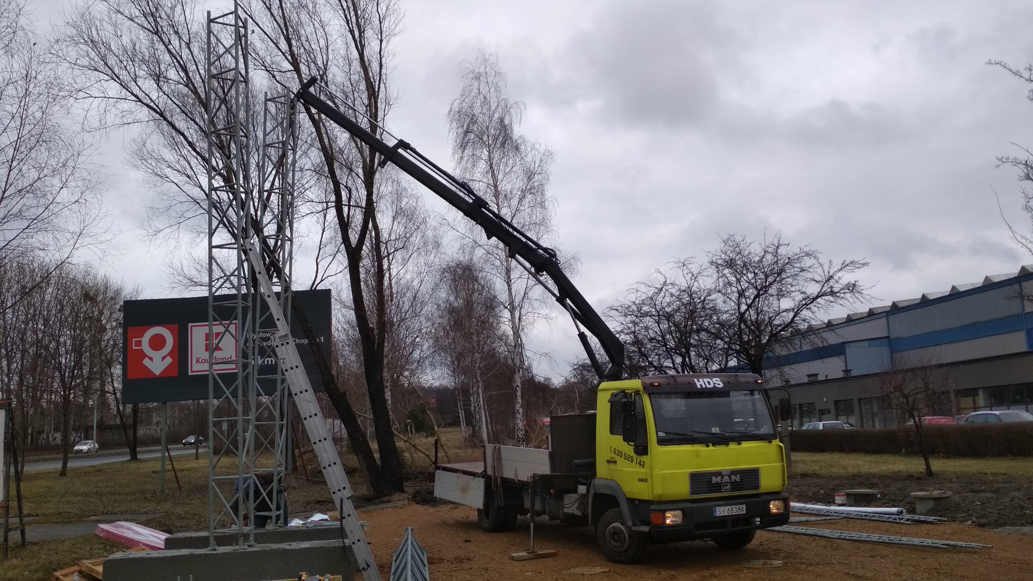 Żółta ciężarówka MAN HDS z wysięgnikiem ustawia metalową konstrukcję pod reklamę Kauflandu na tle pochmurnego nieba i drzew bez liści.