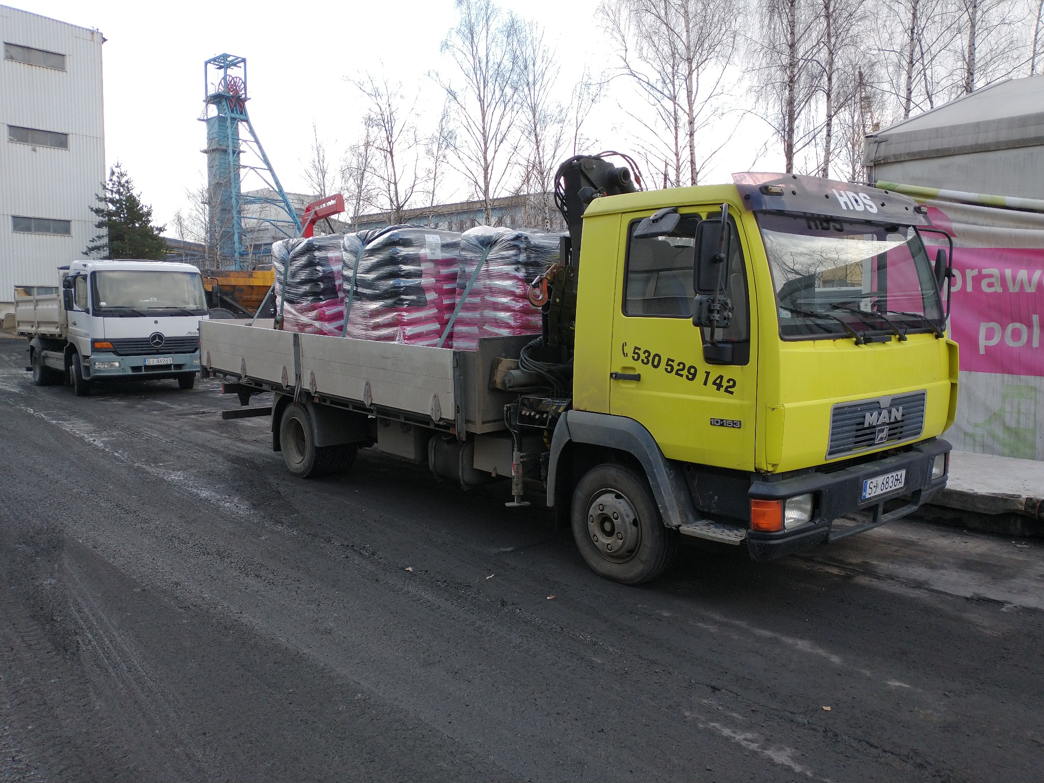 Żółta ciężarówka MAN z HDS załadowana workami na pace, na tle kopalni i innego samochodu ciężarowego.