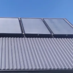 Trzy kolektory słoneczne zamontowane na metalowym dachu z widocznym niebieskim niebem w tle.