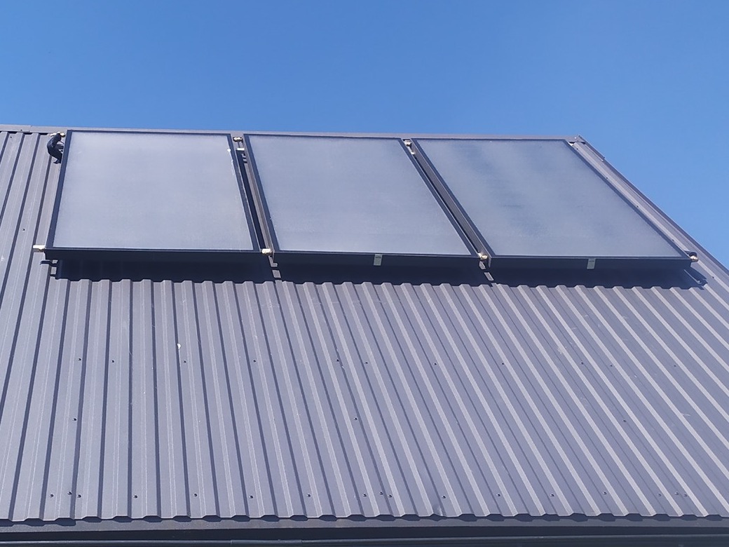 Trzy kolektory słoneczne zamontowane na metalowym dachu z widocznym niebieskim niebem w tle.