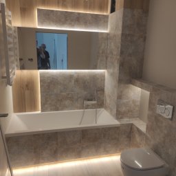 RS Concept Rafał Szlendak - Nowoczesna łazienka z podświetlaną wanną, toaletą podwieszaną i lustrem, wykończona płytkami w odcieniach beżu i brązu z drewnopodobnymi akcentami, odbicie osoby robiącej zdjęcie w lustrze.