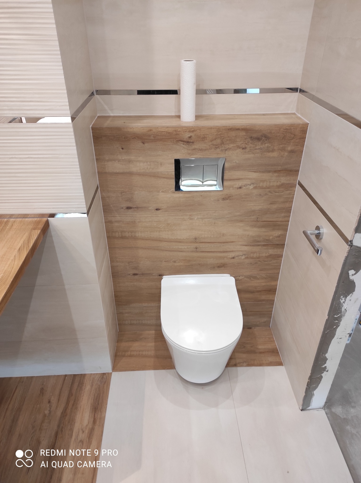 Nowoczesna aranżacja toalety z podwieszaną miską WC, drewnopodobną zabudową stelaża i beżowymi płytkami z metalowymi listwami dekoracyjnymi, widoczny częściowo niezakończony montaż płytek.