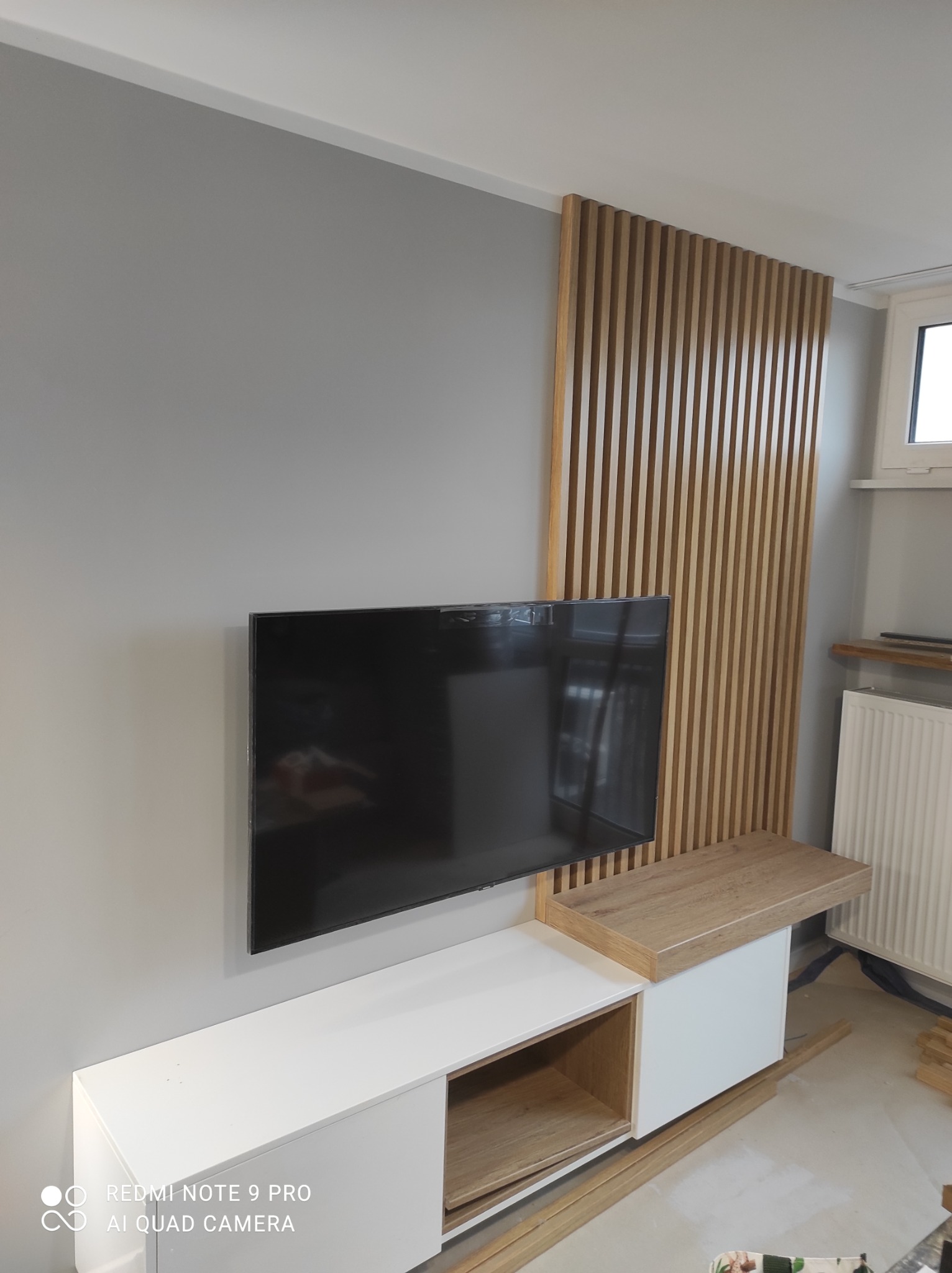 Szara ściana z zamontowanym telewizorem, białą szafką RTV z drewnianymi akcentami i dekoracyjną ścianką z drewnianych listewek, tworzącą nowoczesny wystrój wnętrza.