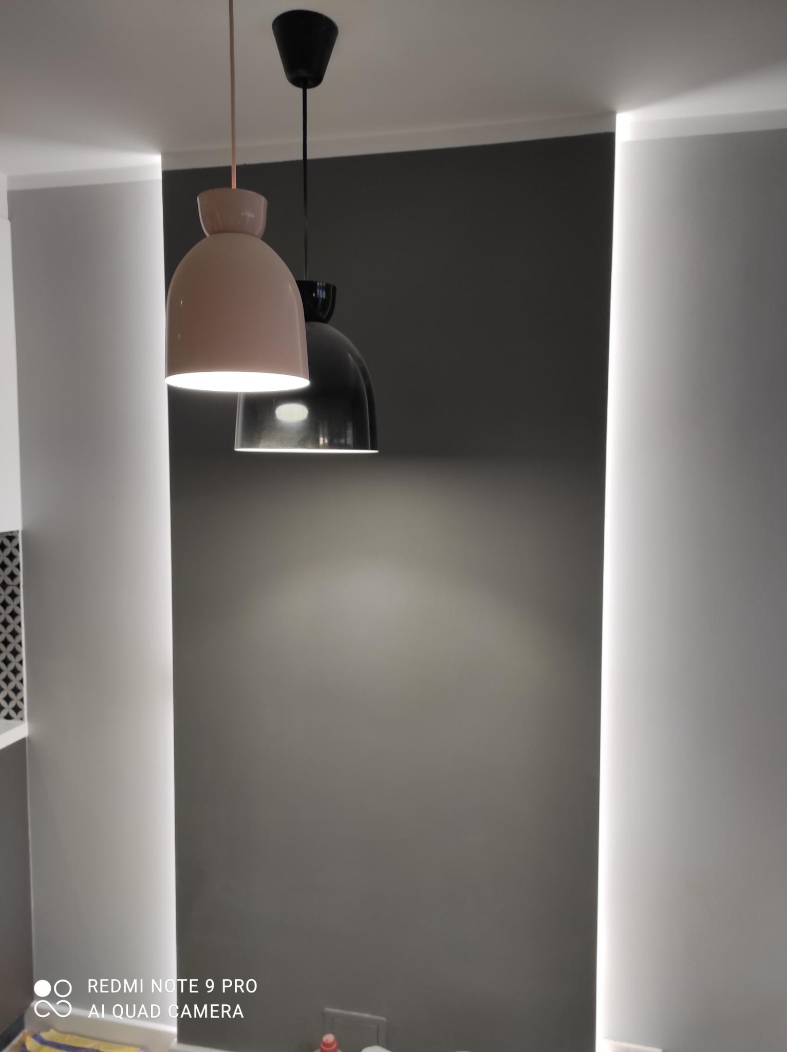 Dwa nowoczesne, wiszące klosze o różnych kształtach i kolorach oświetlają szarą ścianę z wnękami z podświetleniem LED w Grodzisku Mazowieckim.