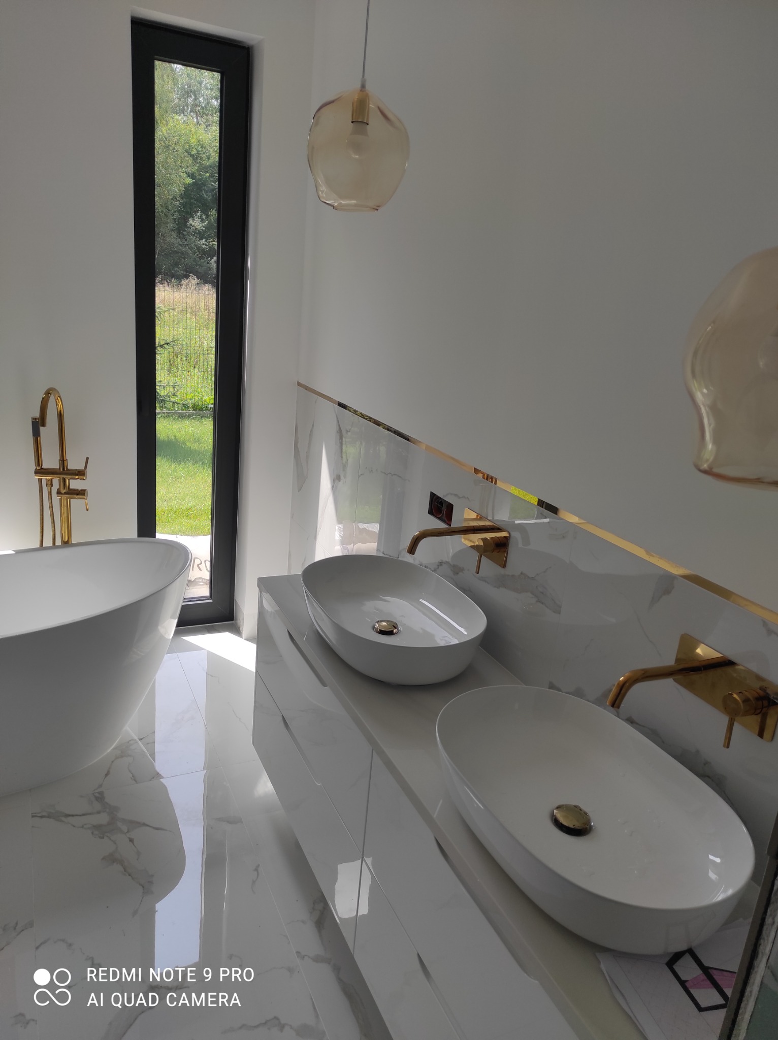 Nowoczesna łazienka z wolnostojącą wanną, dwiema umywalkami nablatowymi z złotą armaturą, białymi płytkami imitującymi marmur i widokiem na zieleń za oknem.