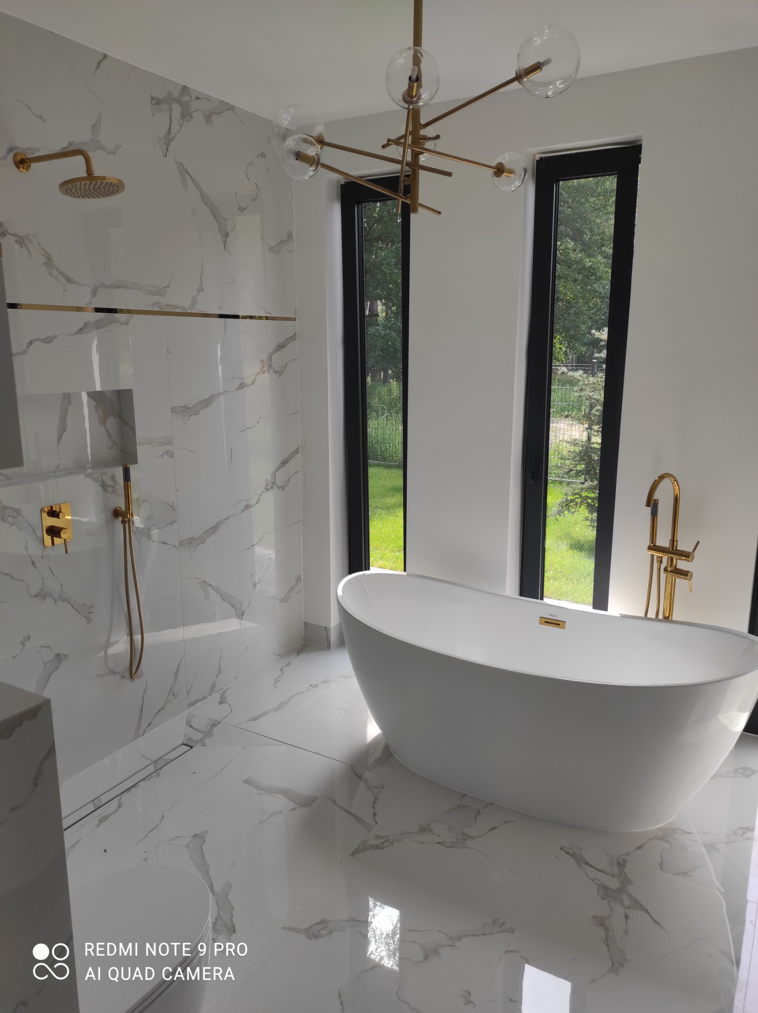 Nowoczesna łazienka z białą wanną wolnostojącą, złotą armaturą i marmurowymi płytkami, widok na zieleń przez okno.