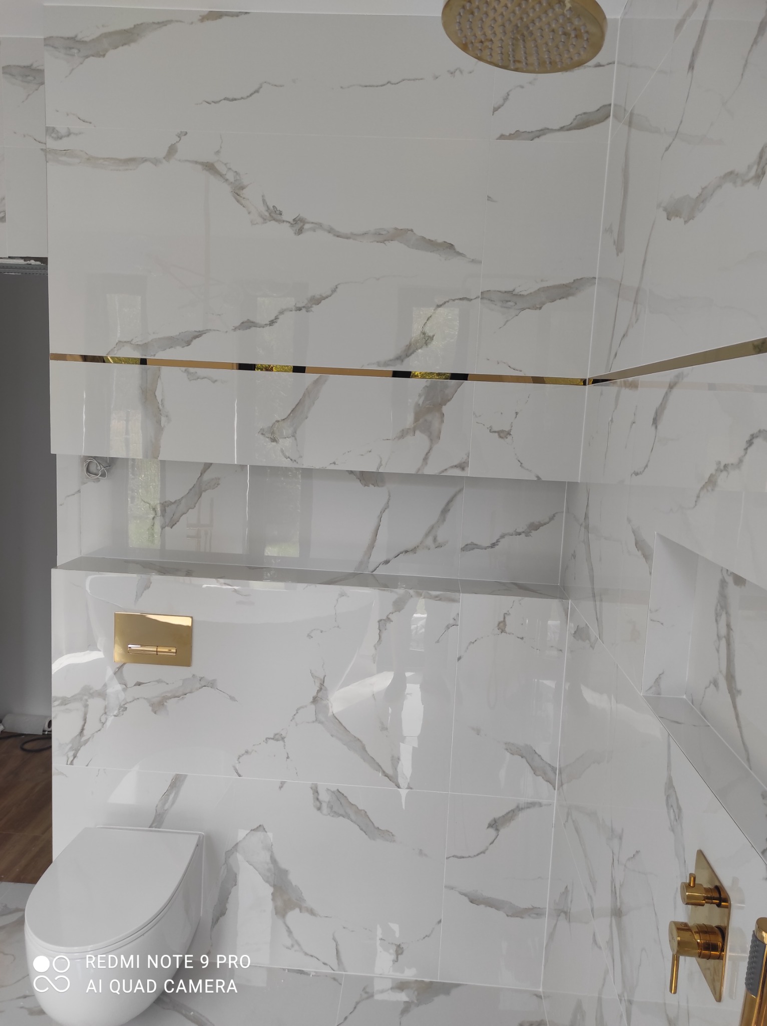 Wykończona łazienka z białymi płytkami imitującymi marmur z złotymi akcentami, widoczna toaleta podwieszana, wnęki w ścianie i złota deszczownica.