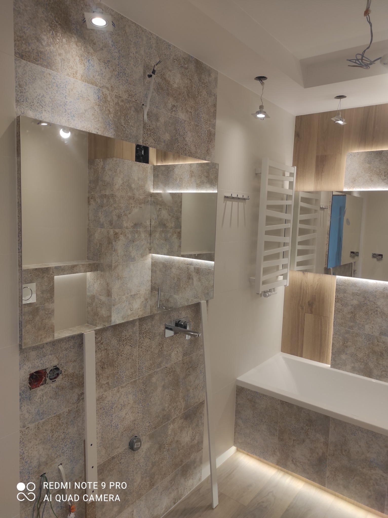 Łazienka w trakcie remontu: częściowo położone płytki, odsłonięte kable elektryczne, zamontowane lustro z podświetleniem, grzejnik drabinkowy i wanna.