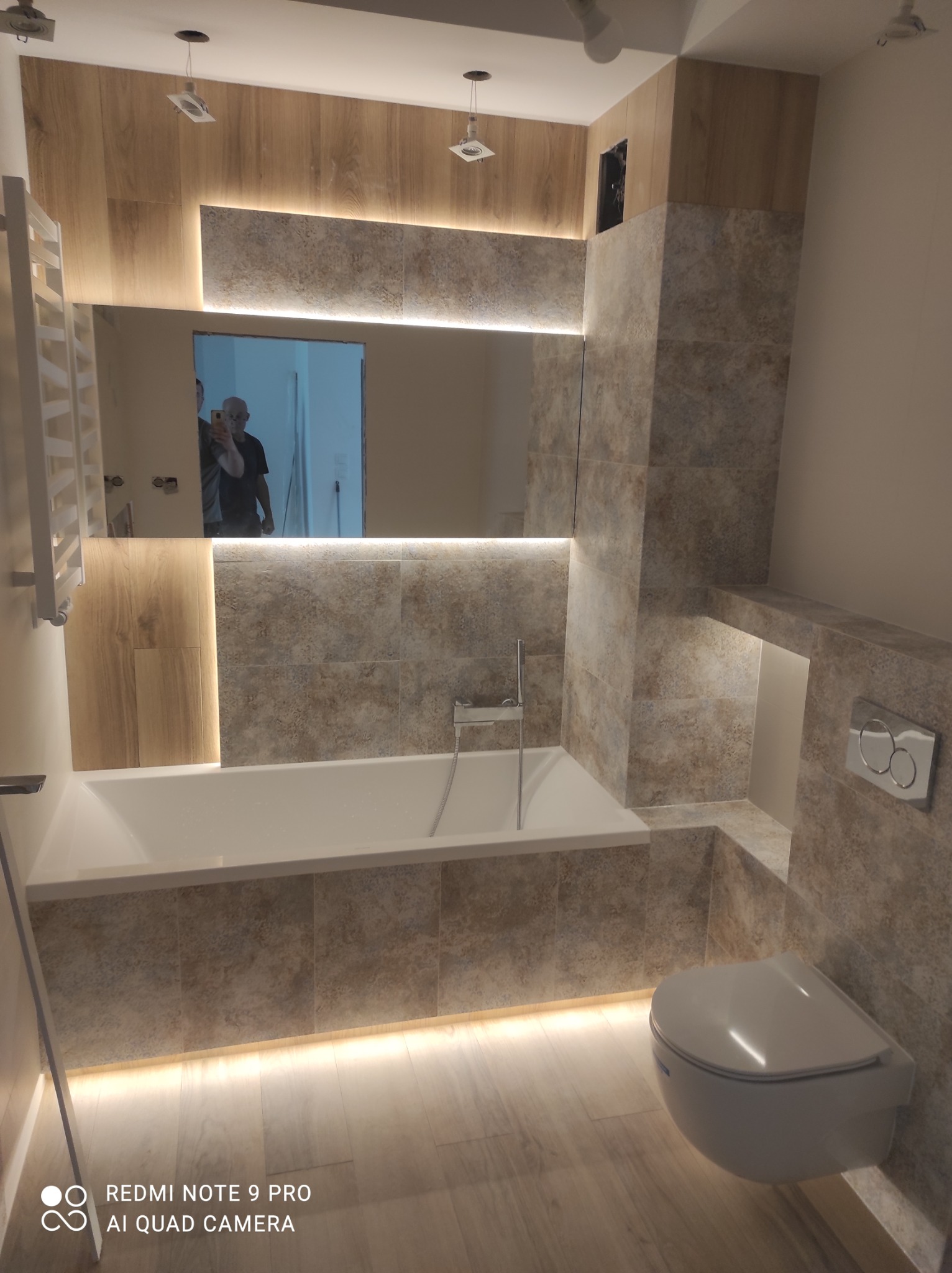 Nowoczesna łazienka z podświetlaną wanną, toaletą podwieszaną i lustrem, wykończona płytkami w odcieniach beżu i brązu z drewnopodobnymi akcentami, odbicie osoby robiącej zdjęcie w lustrze.