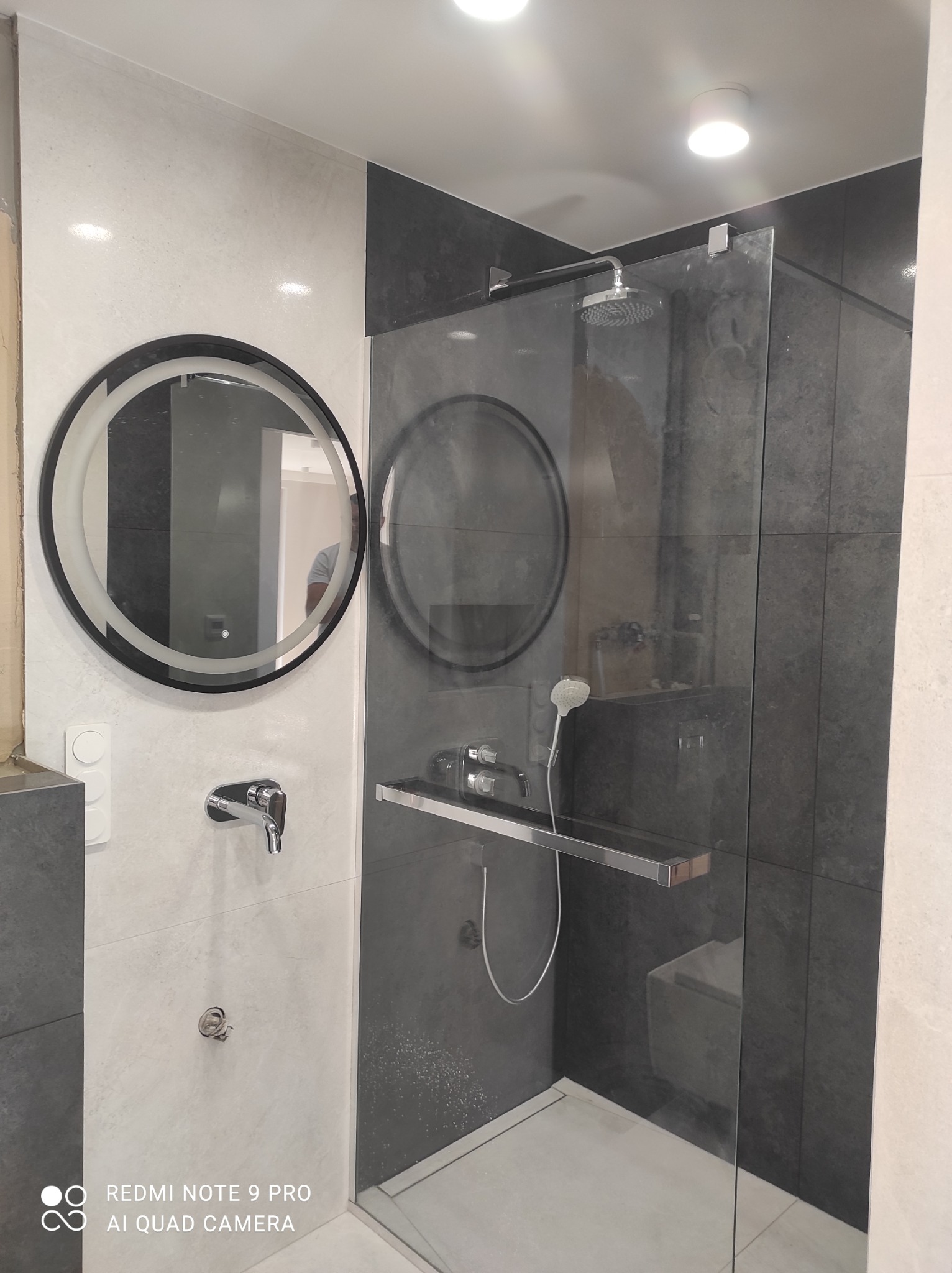 Nowoczesna łazienka z okrągłym lustrem z podświetleniem LED, szklaną kabiną prysznicową z deszczownicą i baterią podtynkową na ścianie wyłożonej jasnymi i ciemnymi płytkami.