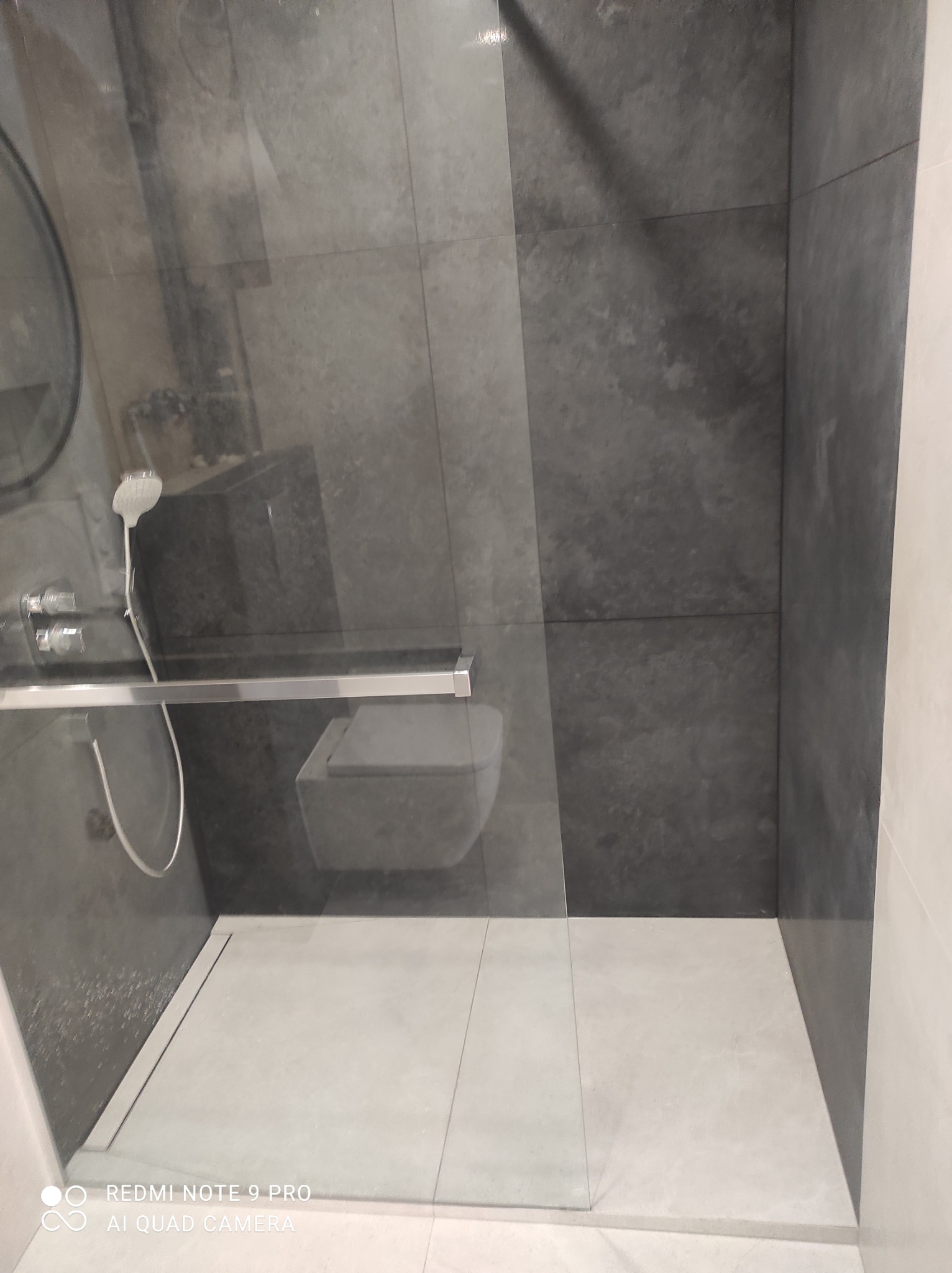 Nowoczesna kabina prysznicowa z szarymi płytkami imitującymi kamień na ścianach i jasnymi płytkami na podłodze, widoczny zestaw prysznicowy i toaleta w tle.