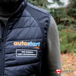 Czarna, pikowana kamizelka robocza z logo firmy 'autostart FHU' i napisem 'Twój dostawca części samochodowych', noszona na swetrze z golfem.