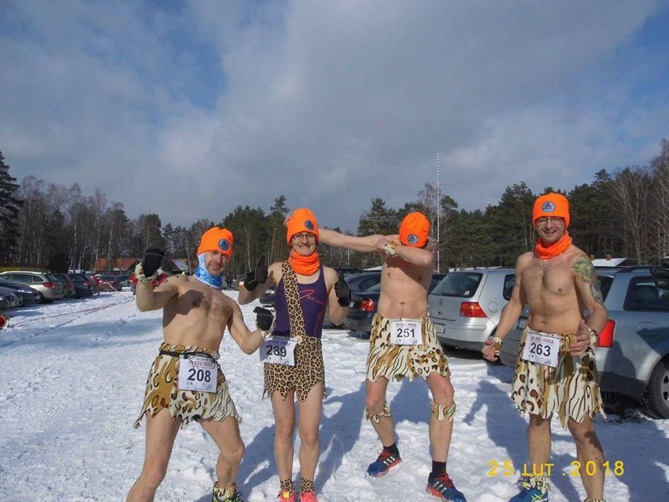 Grupa czterech mężczyzn w zimowej scenerii, ubranych w kostiumy w stylu jaskiniowców z pomarańczowymi czapkami, uczestniczących w biegu, widoczny parking z samochodami w tle.