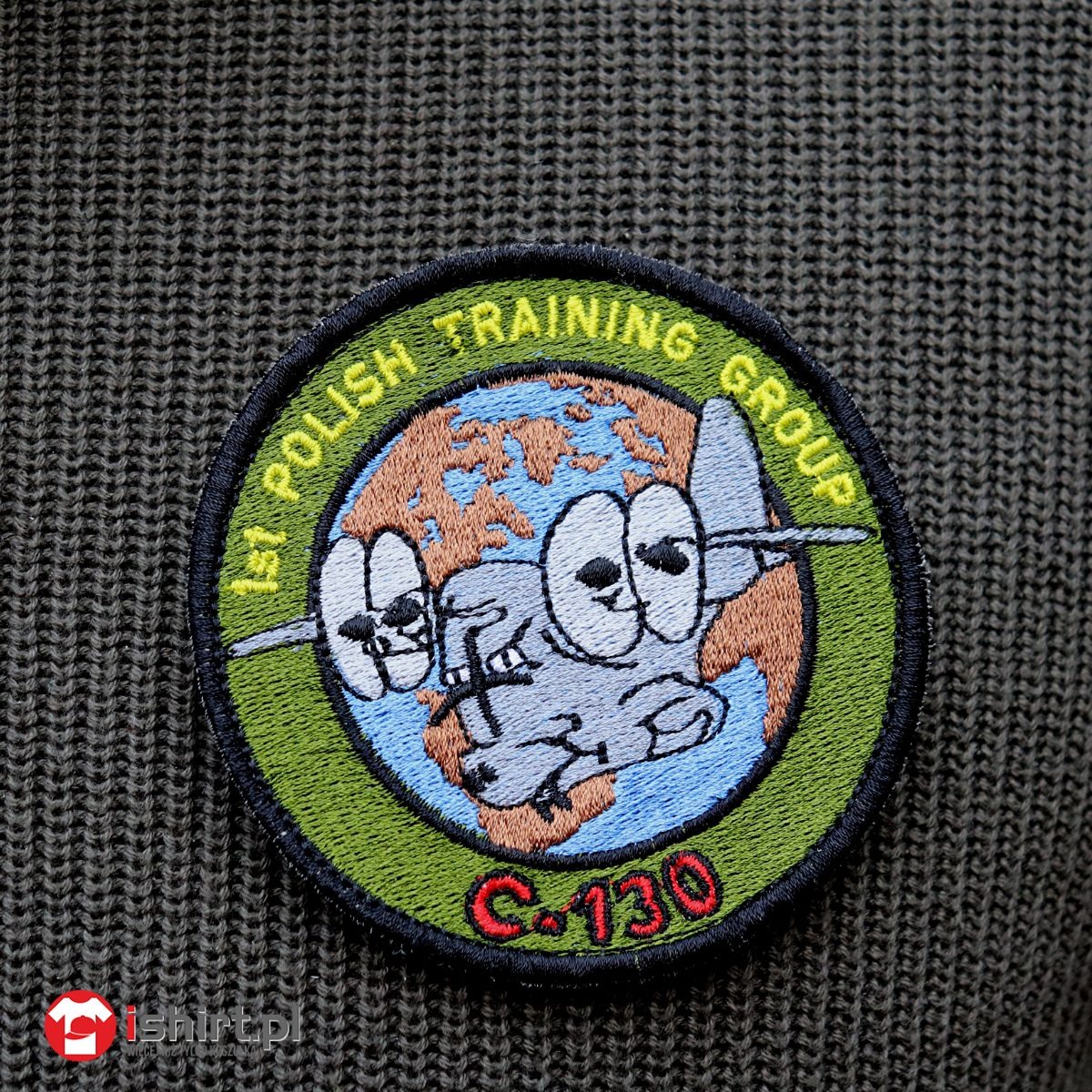 Naszywka haftowana na zielonym swetrze z motywem kreskówkowego samolotu C-130 na tle mapy świata, otoczona napisem '1st Polish Training Group'.