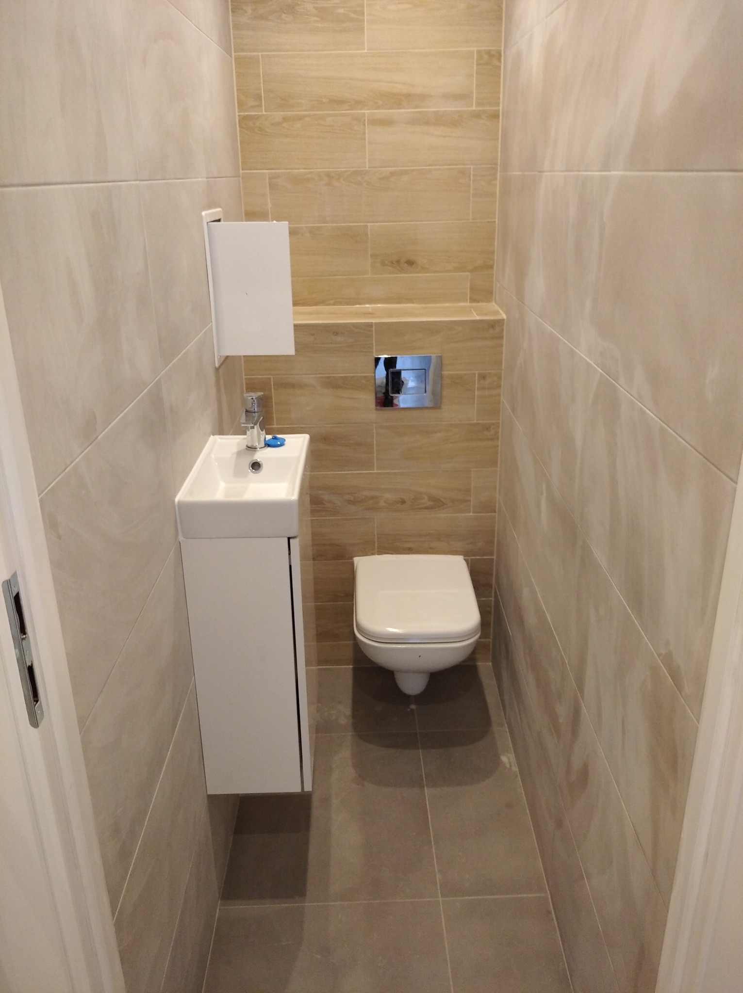 Mała łazienka z beżowymi płytkami imitującymi drewno na ścianach, białą umywalką z szafką, wiszącą toaletą i szarą podłogą z płytek. Widoczny dozownik do mydła przy umywalce.