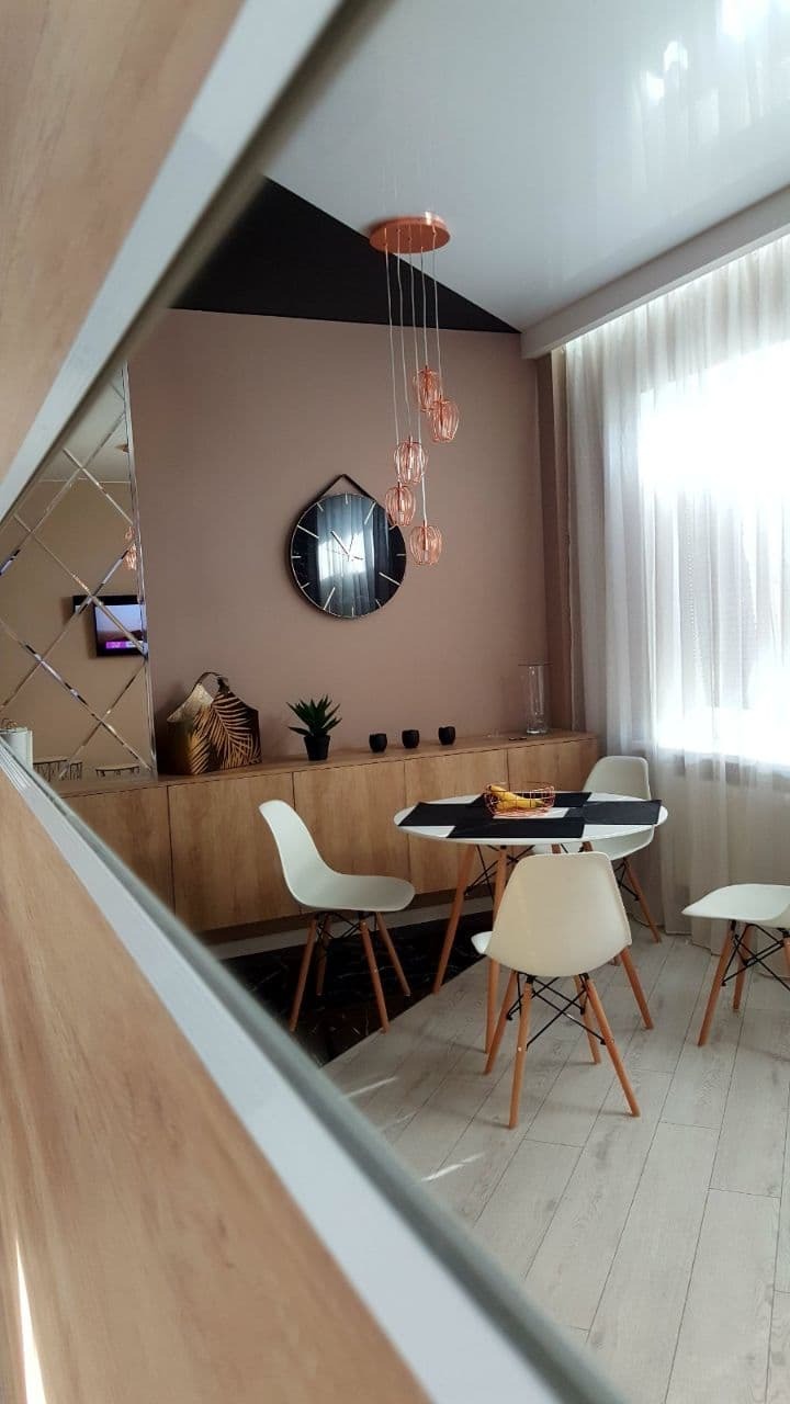 Jasne wnętrze jadalni z okrągłym stołem i czterema krzesłami Eames, oświetlone miedzianą lampą wiszącą, z widocznym fragmentem lustrzanej ściany i dekoracyjną czarną plamą na suficie.