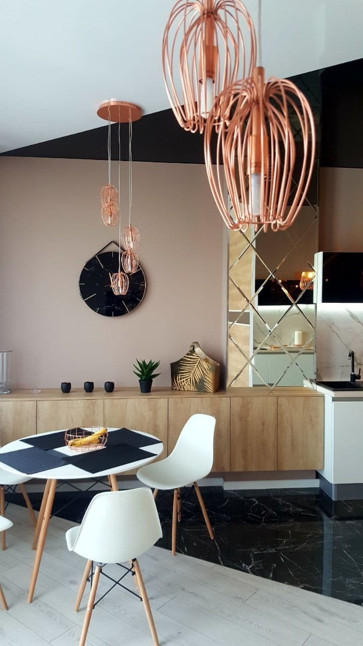 Nowoczesna kuchnia z czarno-białą podłogą imitującą marmur, drewnianymi szafkami, okrągłym stołem z białymi krzesłami i miedzianymi lampami wiszącymi.
