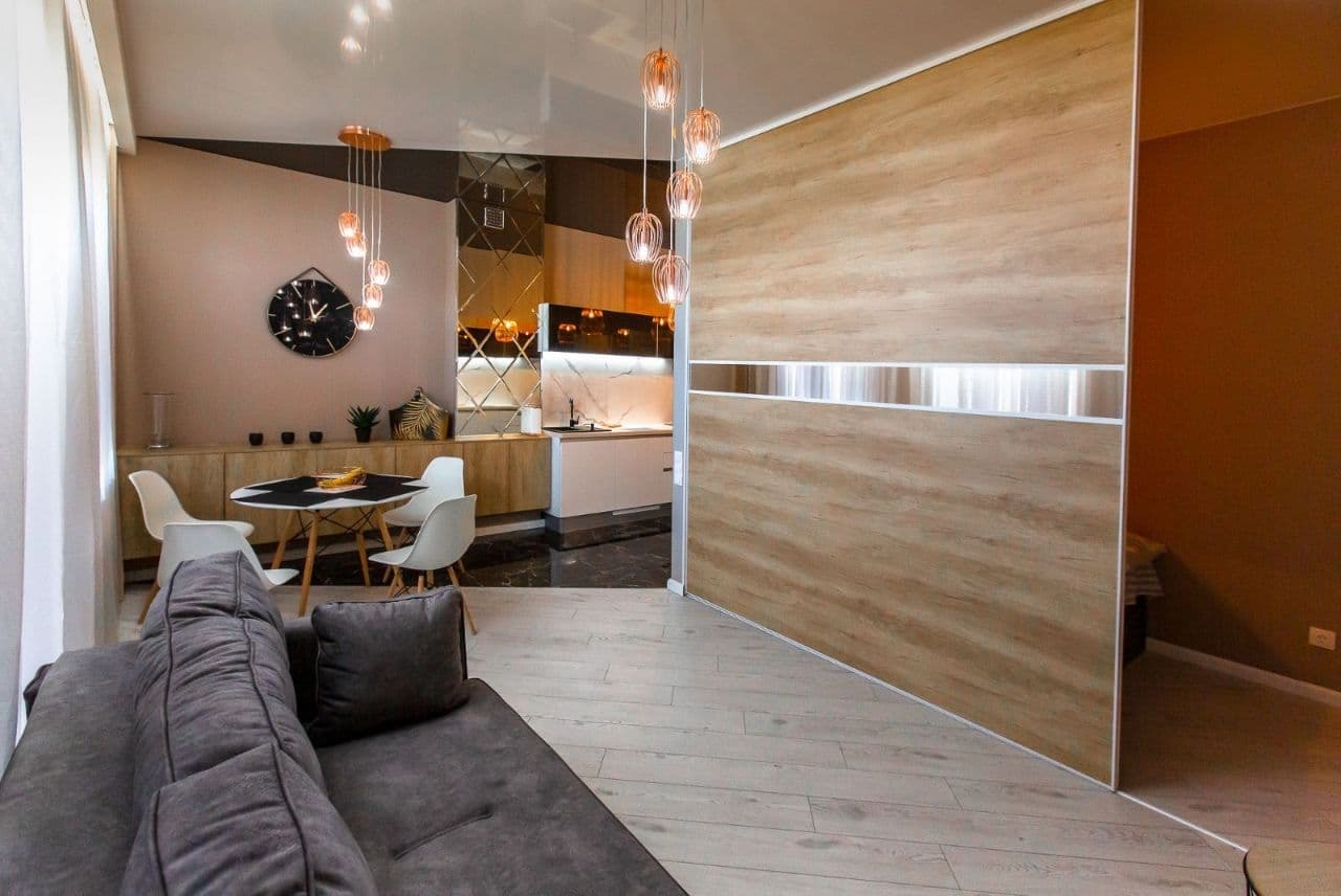 Nowoczesne wnętrze mieszkania z drewnianą ścianą działową z poziomym pasem lustra, widok na kuchnię z marmurowym blatem i jadalnię z okrągłym stołem oraz szarą sofą.